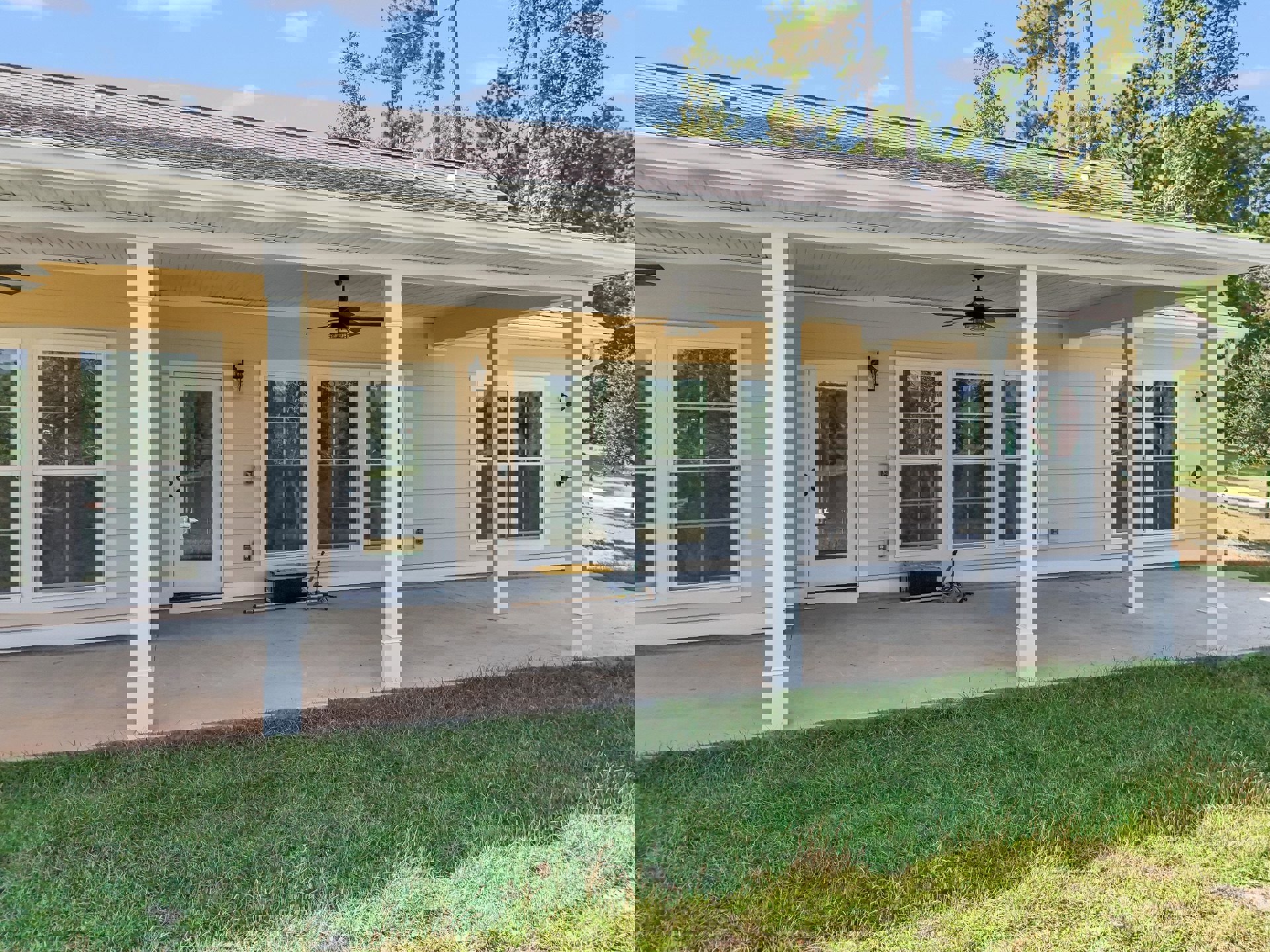 White siding house with covered porch, ceiling fan, concrete patio, and green lawn; glass door reveals lake and boat view, window faces leafy tree, black and yellow storage box