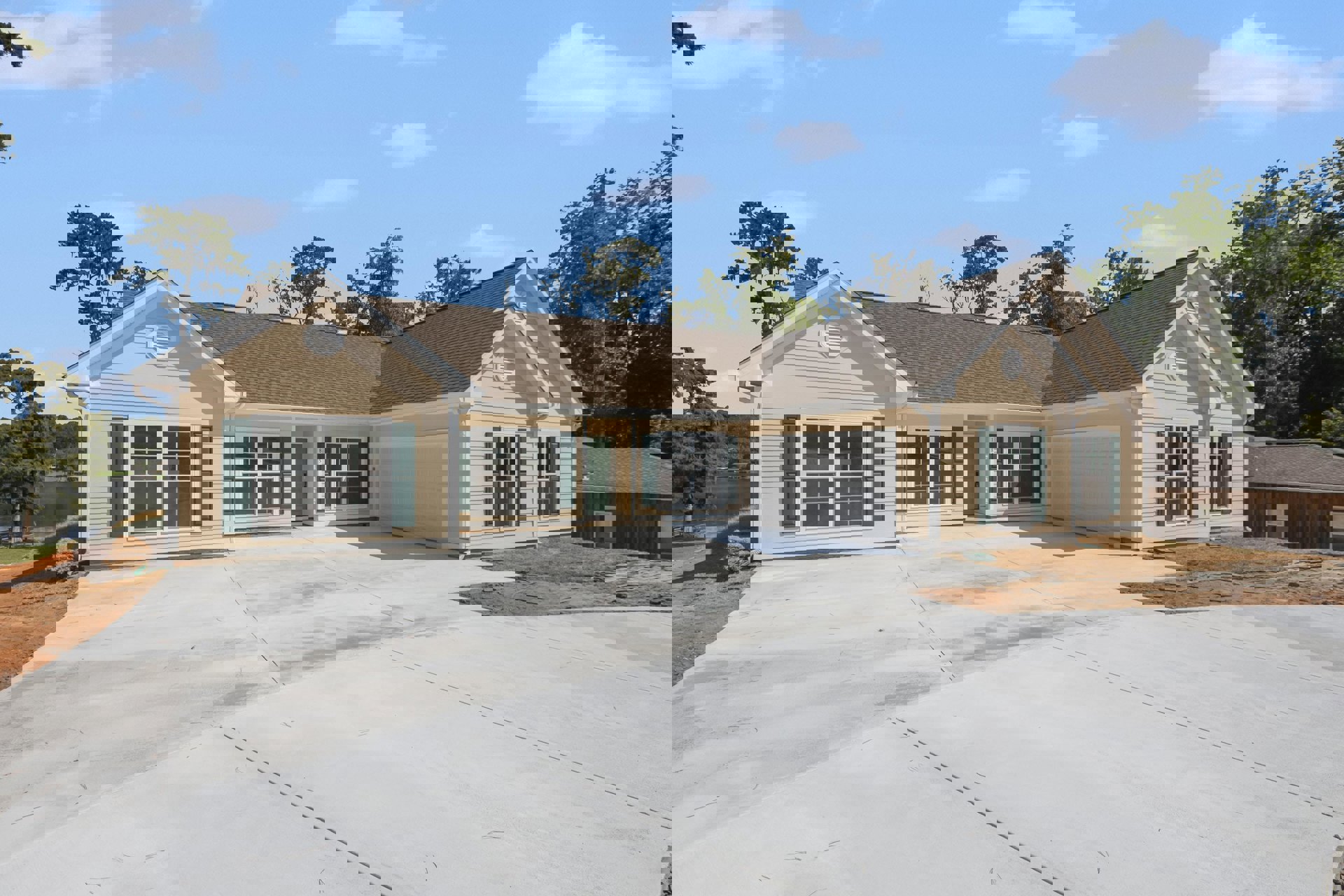 Two-story house with gray siding, green shuttered windows, attached garage, paved driveway, white wall vent, and trees behind the roof under a cloudy sky