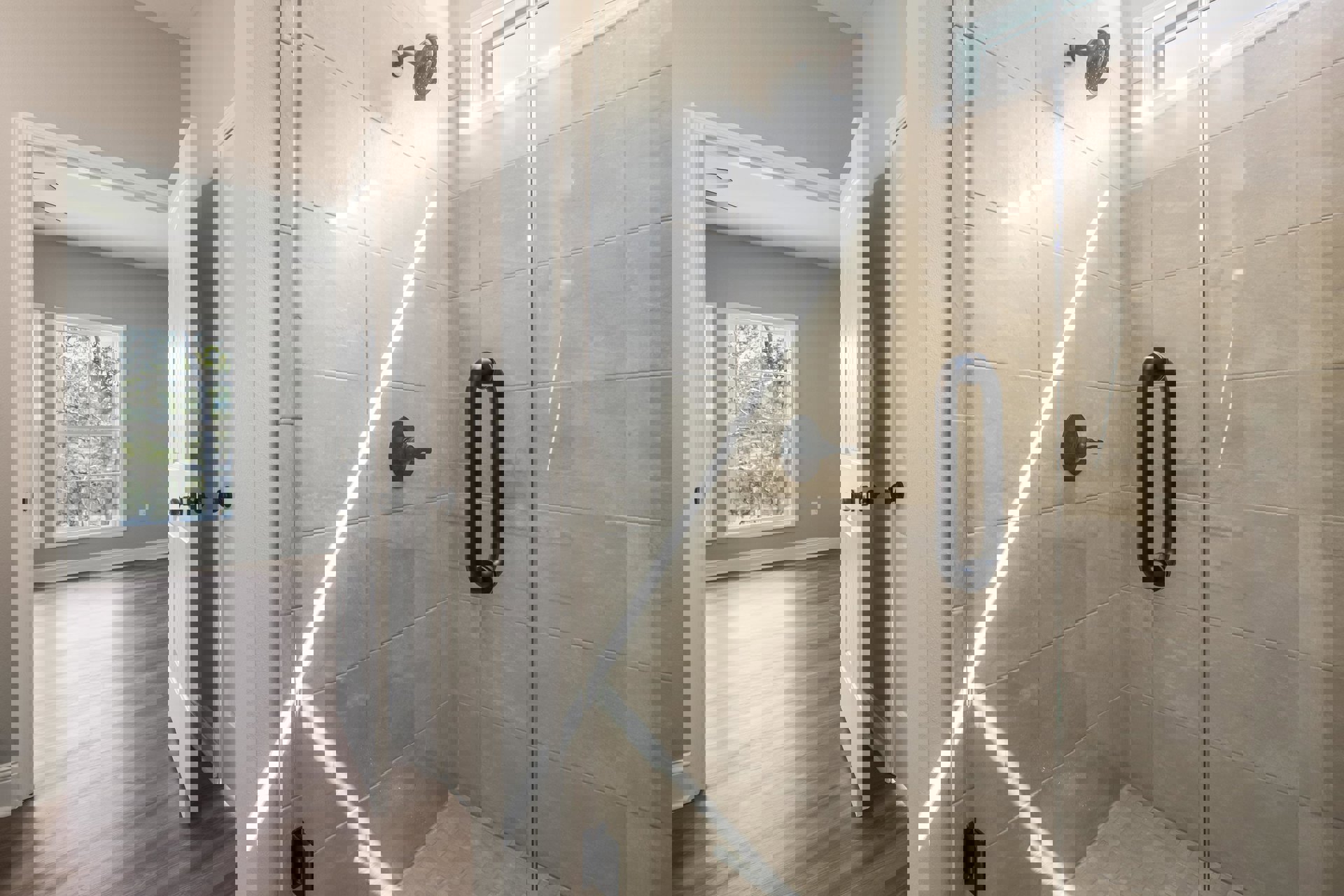 Walk-in shower with clear glass door, chrome shower head mounted on tiled wall, black metal door knob, wood flooring, open white door, window overlooking trees.