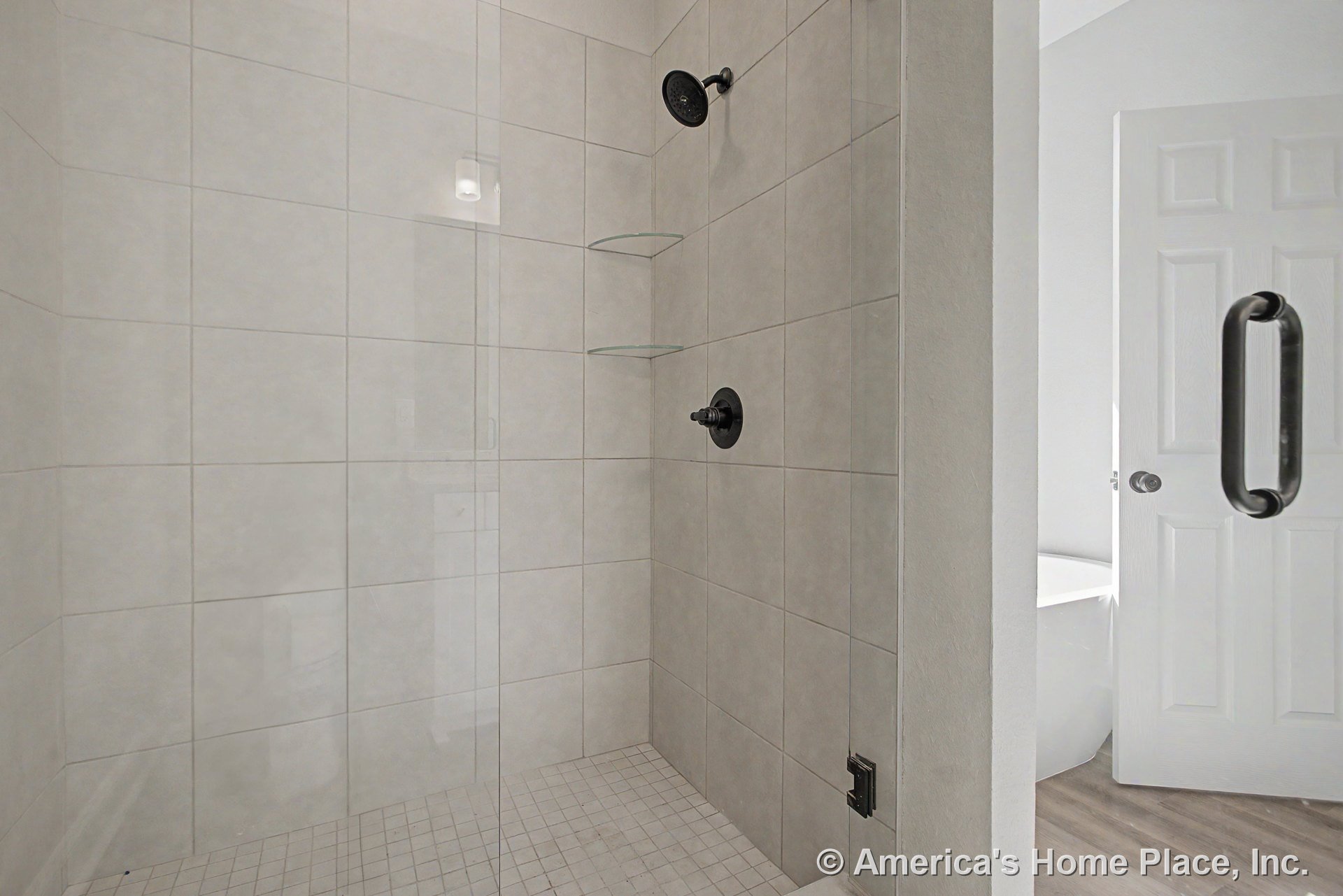 Walk-in shower with large light-colored wall tile, glass shower door, glass corner shelves, matte black shower fixtures, and white paneled door.