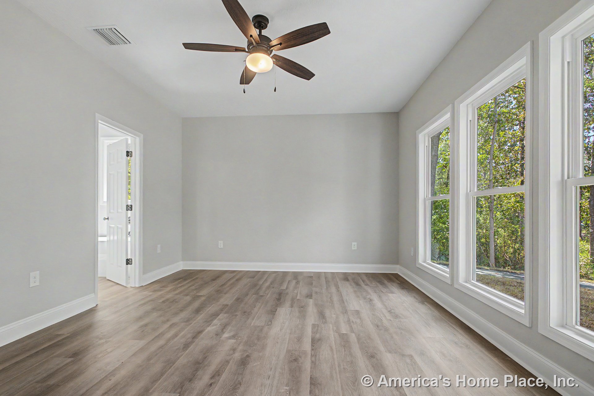 Large triple windows with white trim fill the bedroom with natural light, highlighting light gray walls, modern wood-look vinyl plank flooring, white baseboards, and a ceiling fan