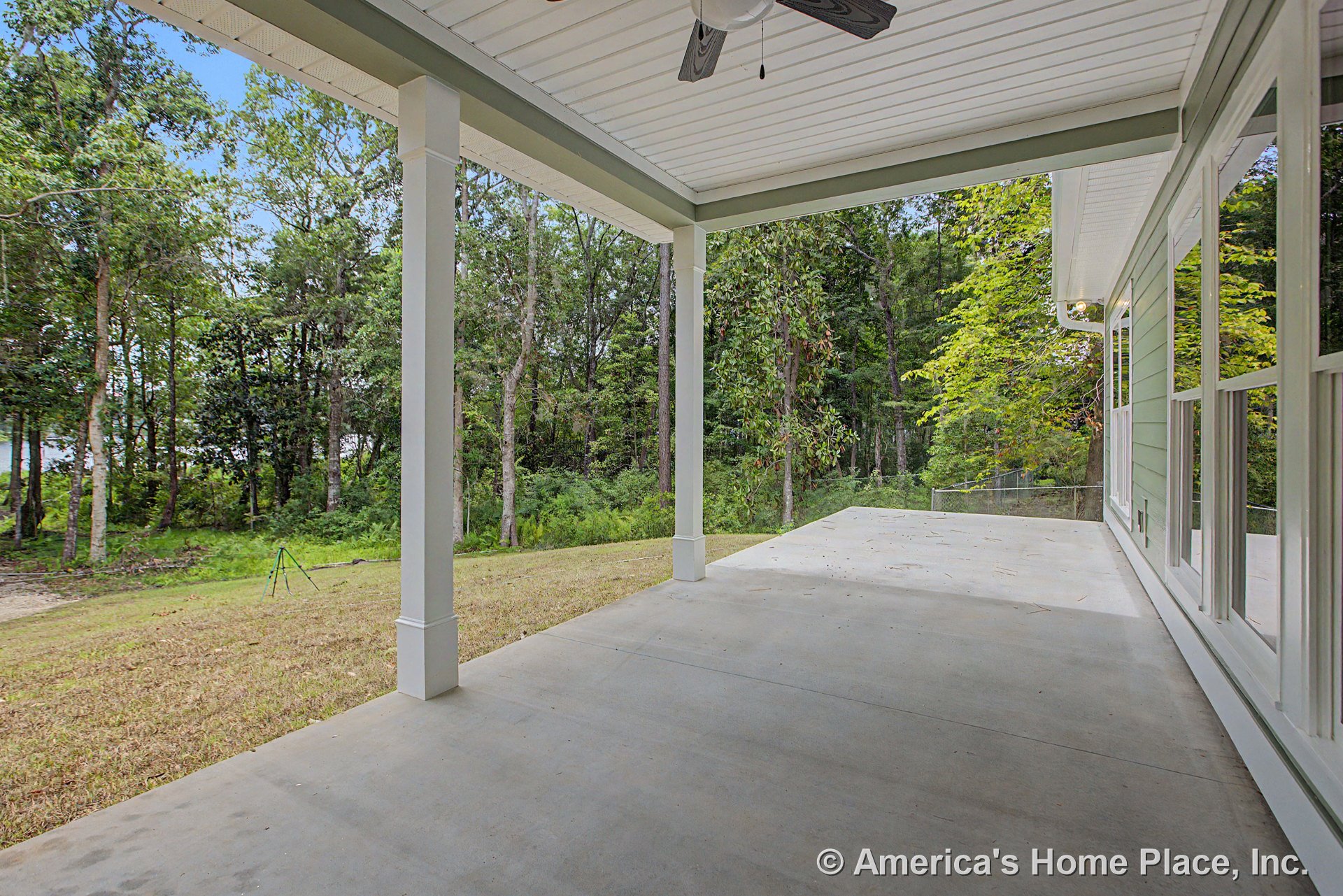 Covered concrete patio with white column supports, ceiling fan fixture, and porch ceiling, facing large exterior windows and green siding, overlooking wooded backyard.