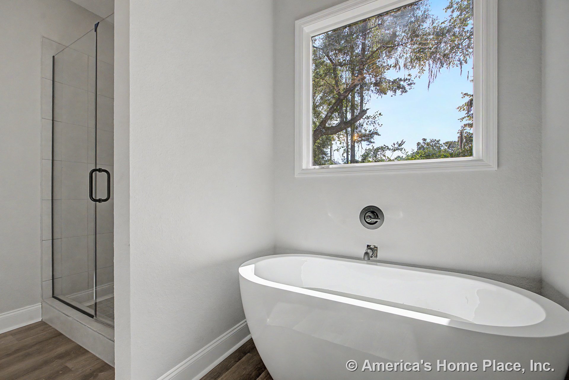 Freestanding soaking tub positioned beneath a large window with decorative trim, glass-enclosed shower featuring tile walls, wood-look flooring, white painted walls, and