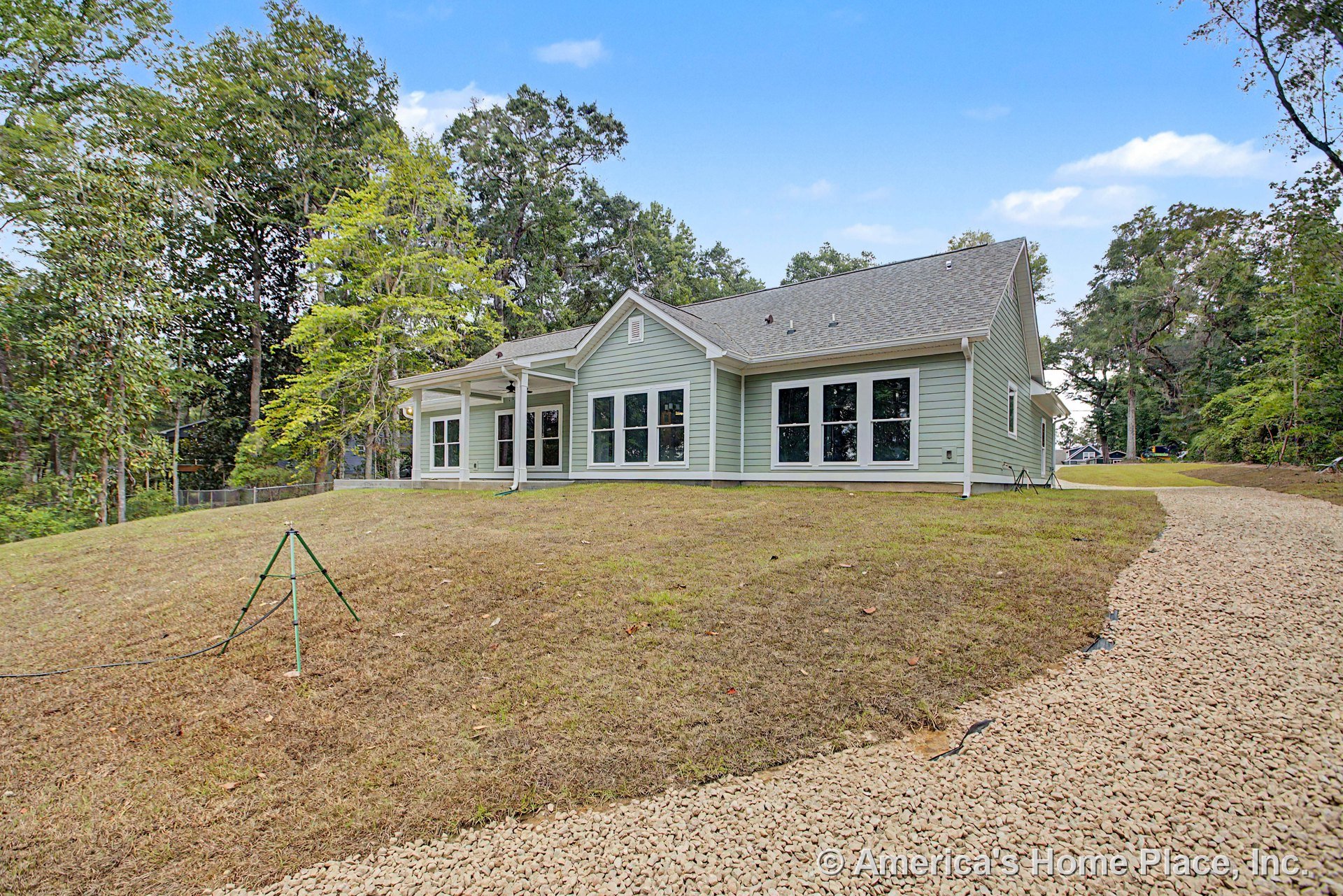 Light green horizontal lap siding with white trim accents, multiple large windows, covered rear porch supported by columns, gray shingle roof, and gravel driveway at rear