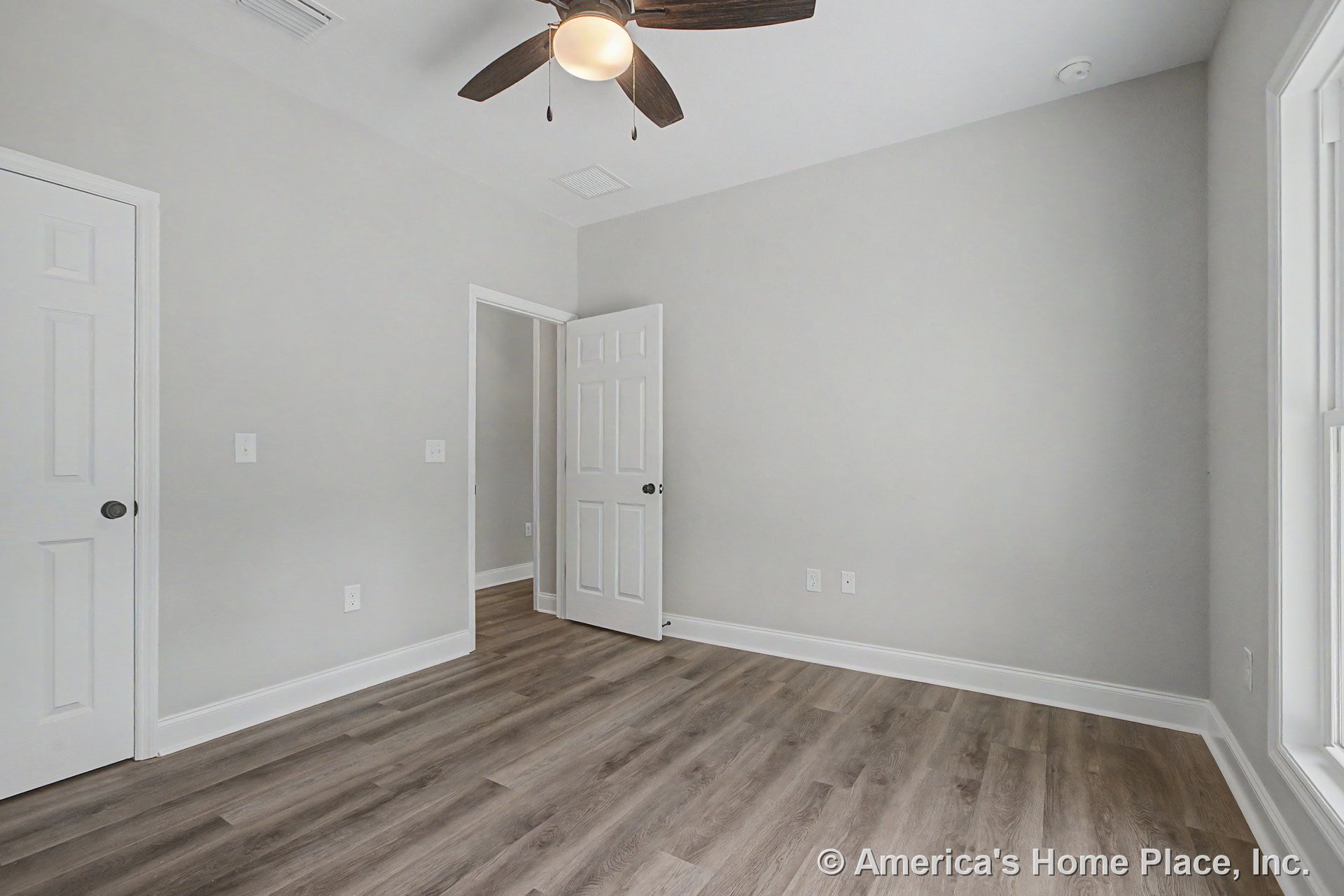 Bedroom with light gray walls, wood-look vinyl plank flooring, white trim and baseboards, paneled interior door, large window, and ceiling fan with integrated light fixture.