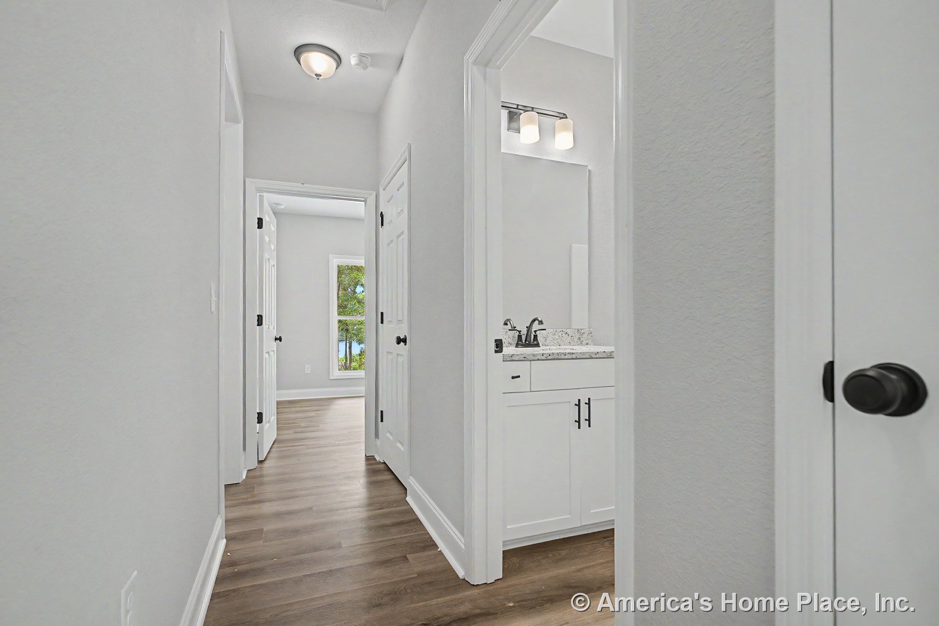 Hallway with wood-look flooring and white trim leading to a bathroom featuring a granite countertop, white shaker-style cabinetry, modern light fixture above the mirror, black
