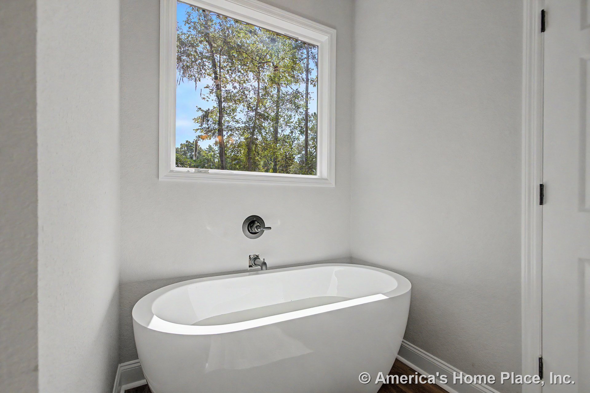 Freestanding bathtub beneath expansive window, light gray walls with white trim, wood-look flooring, modern tub faucet, minimalist bathroom design with abundant natural light.