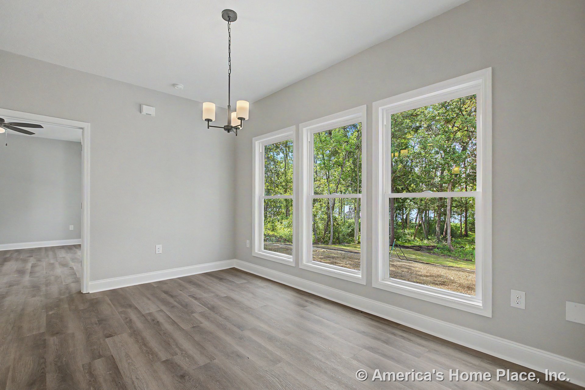 Dining area with three large picture windows, modern chandelier, light gray painted walls, white baseboard trim, wood-look vinyl plank flooring, and open doorway leading to