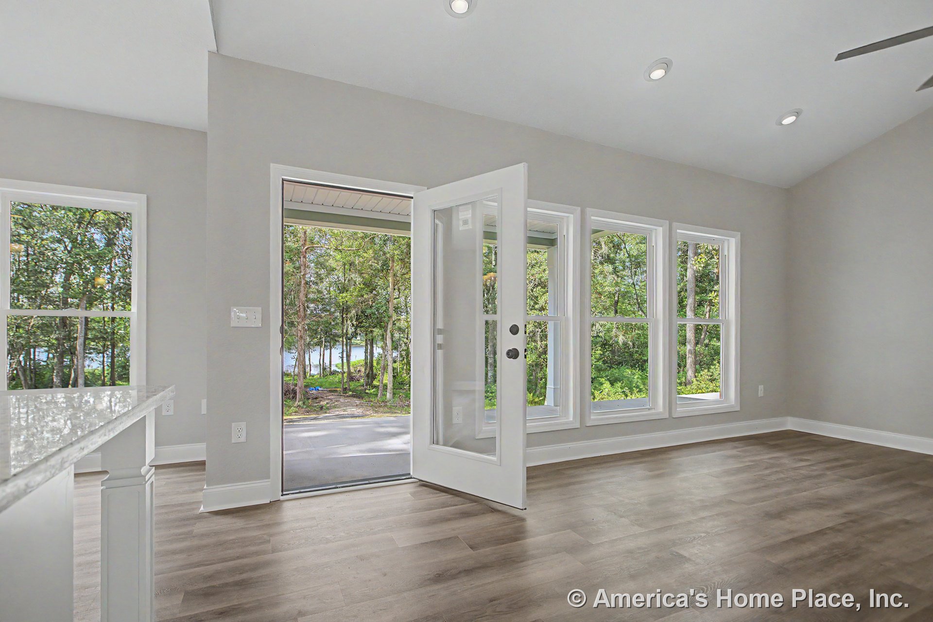 Wide plank wood flooring, multiple large windows with white trim, glass-paneled exterior door, recessed ceiling lights, neutral wall paint, and white baseboards in an open living