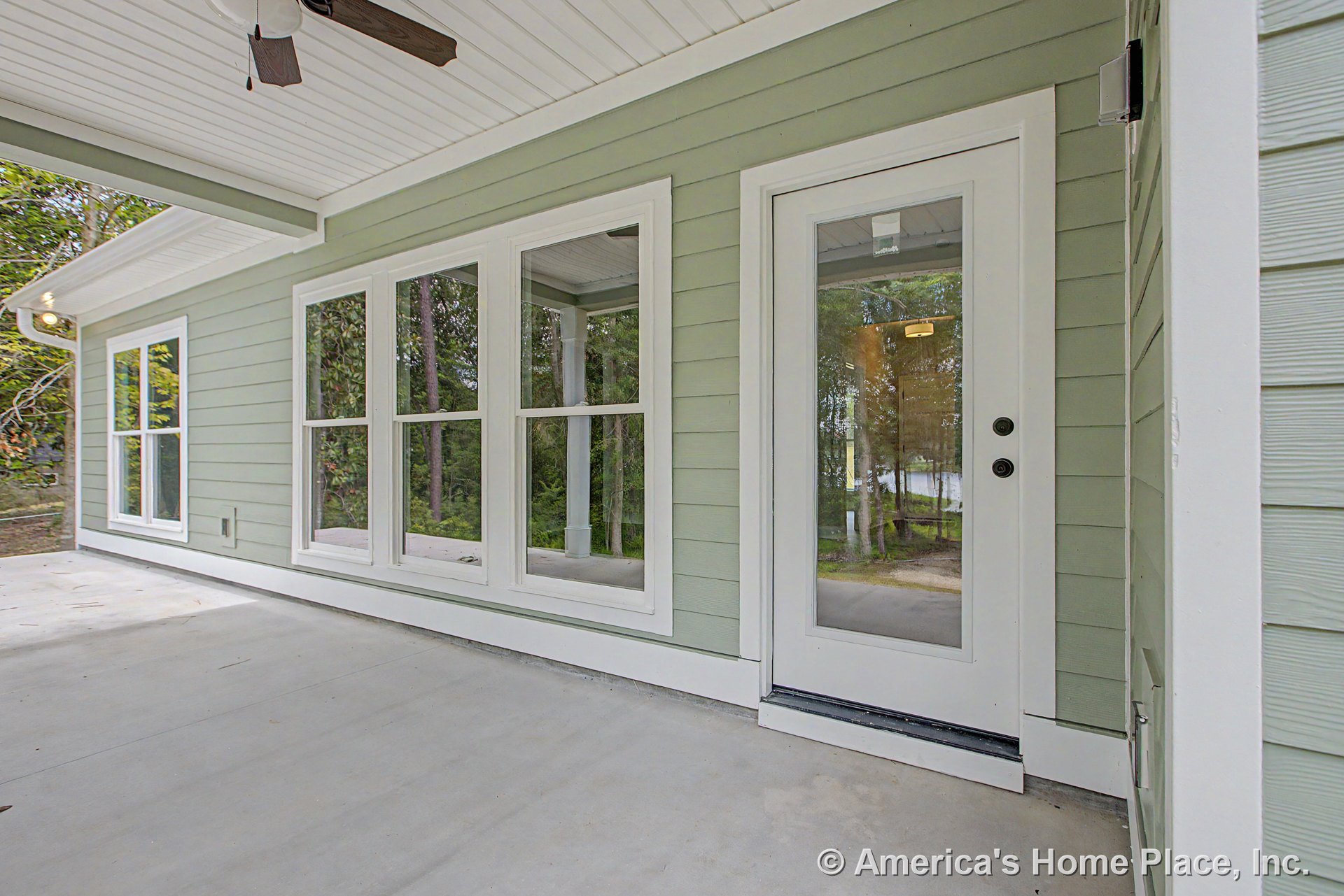 Covered porch with light green horizontal siding, white trim, multiple large white-framed windows, glass exterior door, concrete floor, and ceiling fan.
