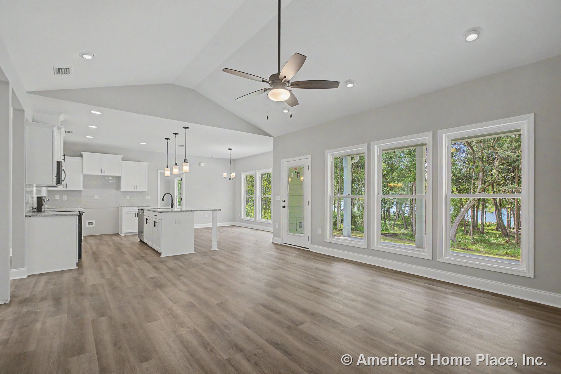 Open-concept living area with vaulted ceiling, recessed lighting, large windows facing wooded view, modern kitchen featuring white cabinetry, granite countertops, central island