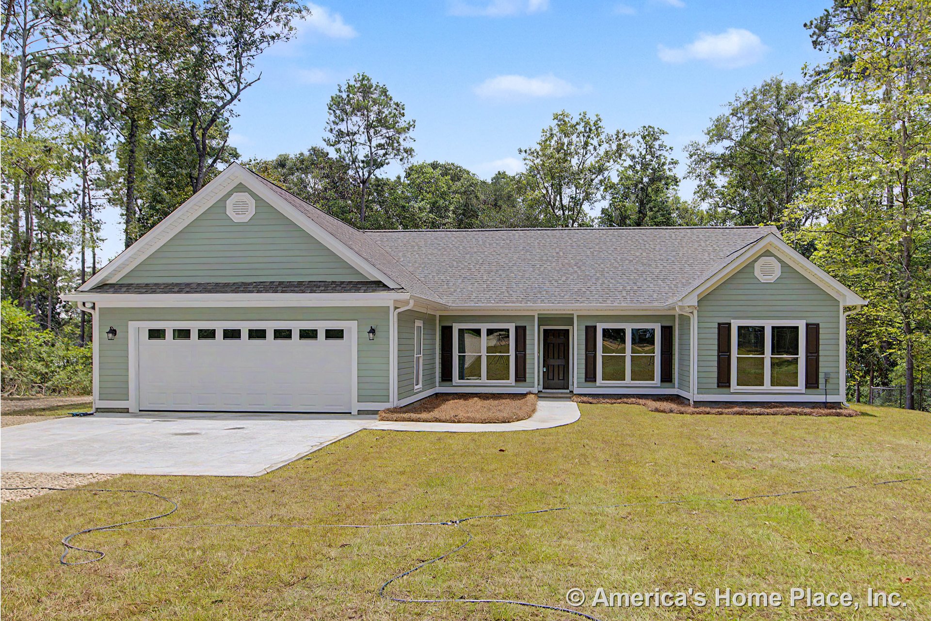 Light green siding with white trim, covered front porch, attached two-car garage with windows, large front windows, gray shingle roof, exterior wall lanterns, paved driveway.