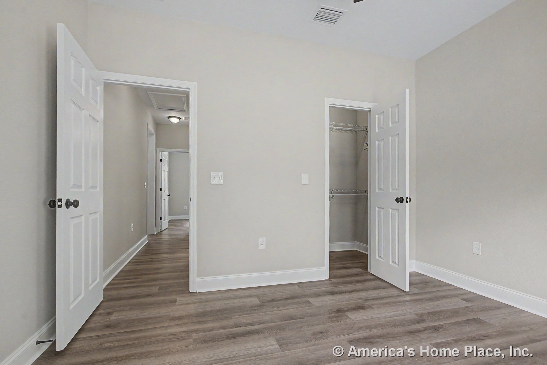 Bedroom with light gray walls, wood-look vinyl plank flooring, white baseboards and trim, open closet featuring wire shelving, white paneled doors, and adjacent hallway with