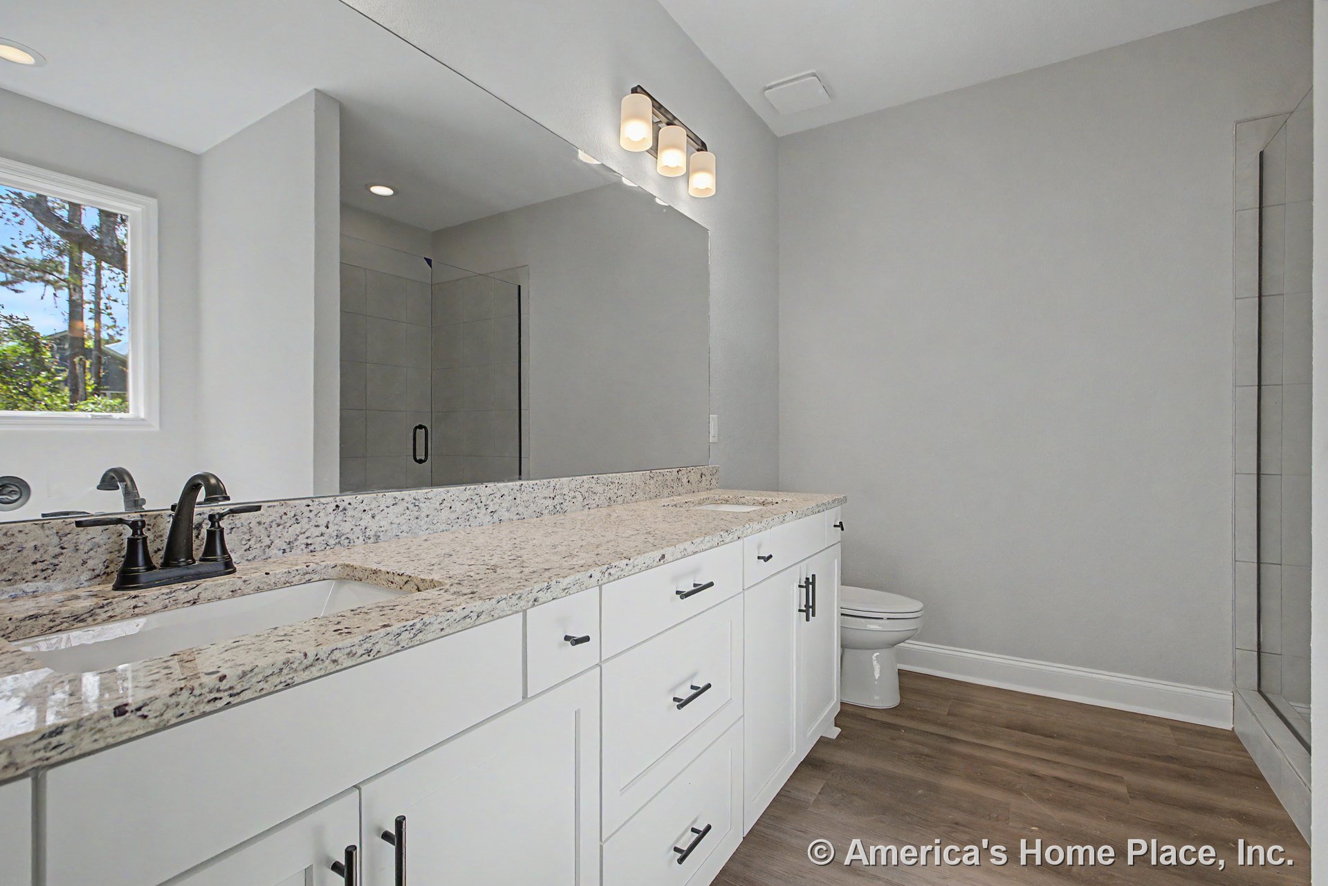 Granite vanity top with single matte black faucet, white shaker cabinets, glass-enclosed tile shower, wood-look flooring, large wall mirror, modern vanity lighting, window, and