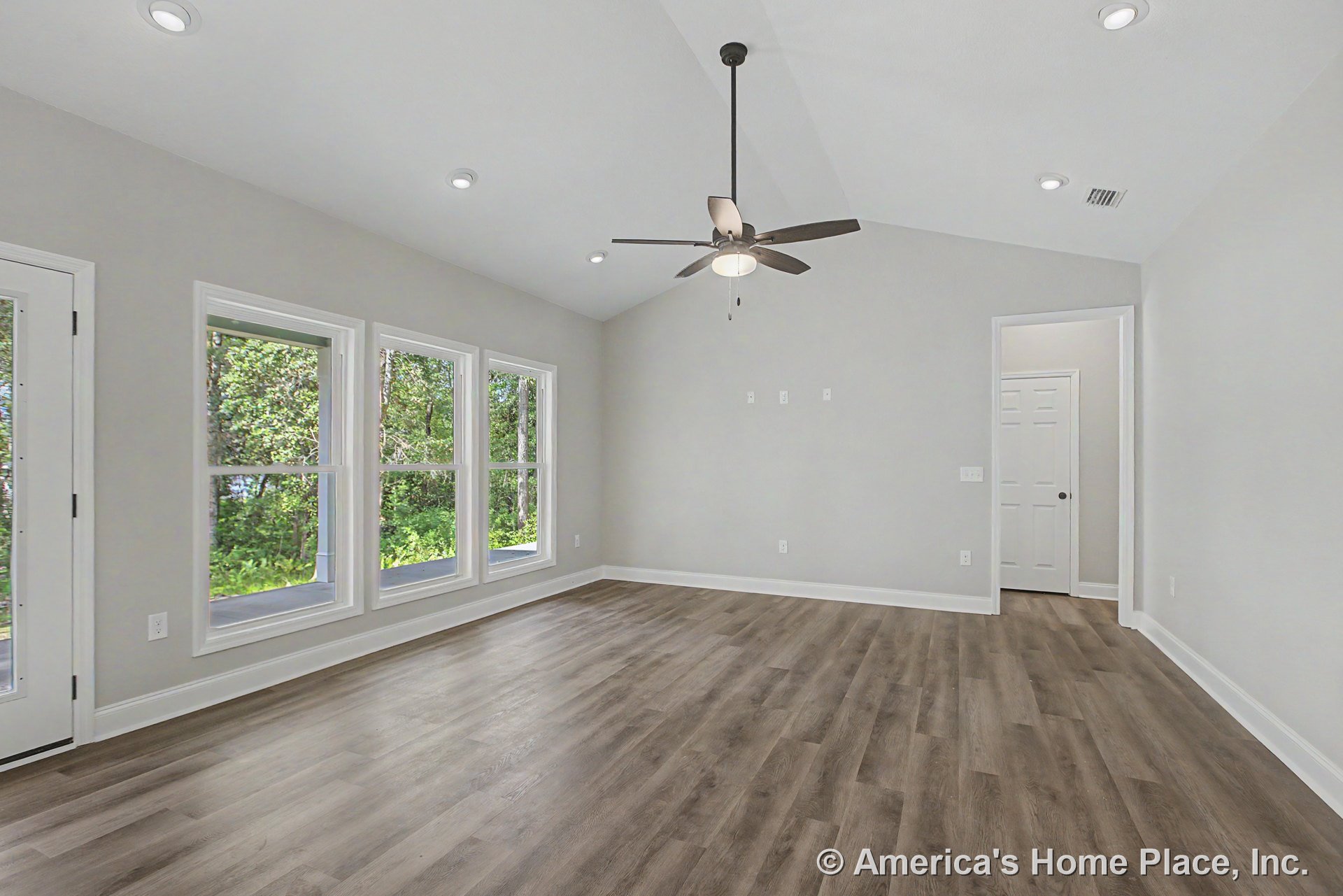 Vaulted ceiling living room with three large windows, recessed ceiling lights, ceiling fan with light, wood-look plank flooring, white trim and baseboards, neutral wall color, and
