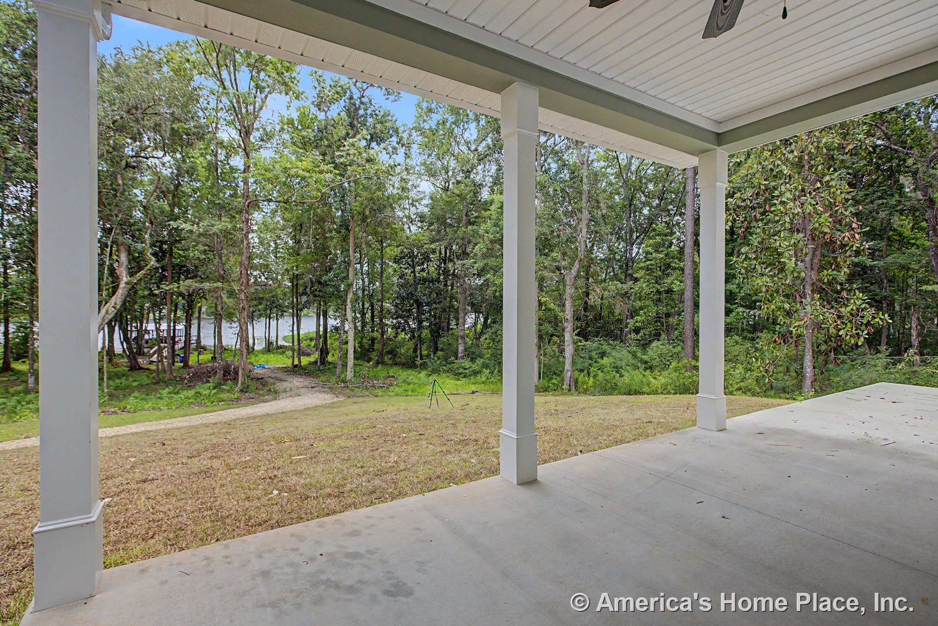 Covered concrete porch with white square columns, trimmed posts, ceiling fan fixture, and overhead ceiling, overlooking wooded backyard and lake through trees.