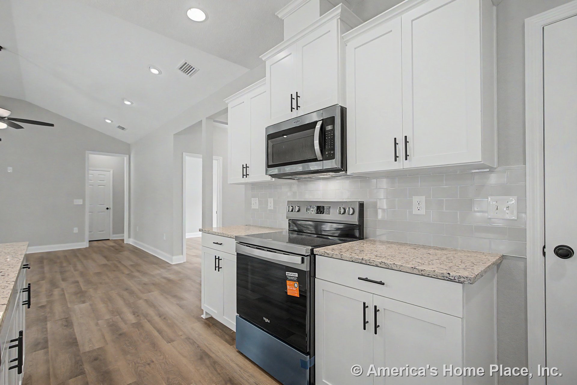 White shaker cabinets with granite countertops, subway tile backsplash, stainless steel range and microwave, recessed ceiling lights, and wood-look flooring in a modern kitchen.