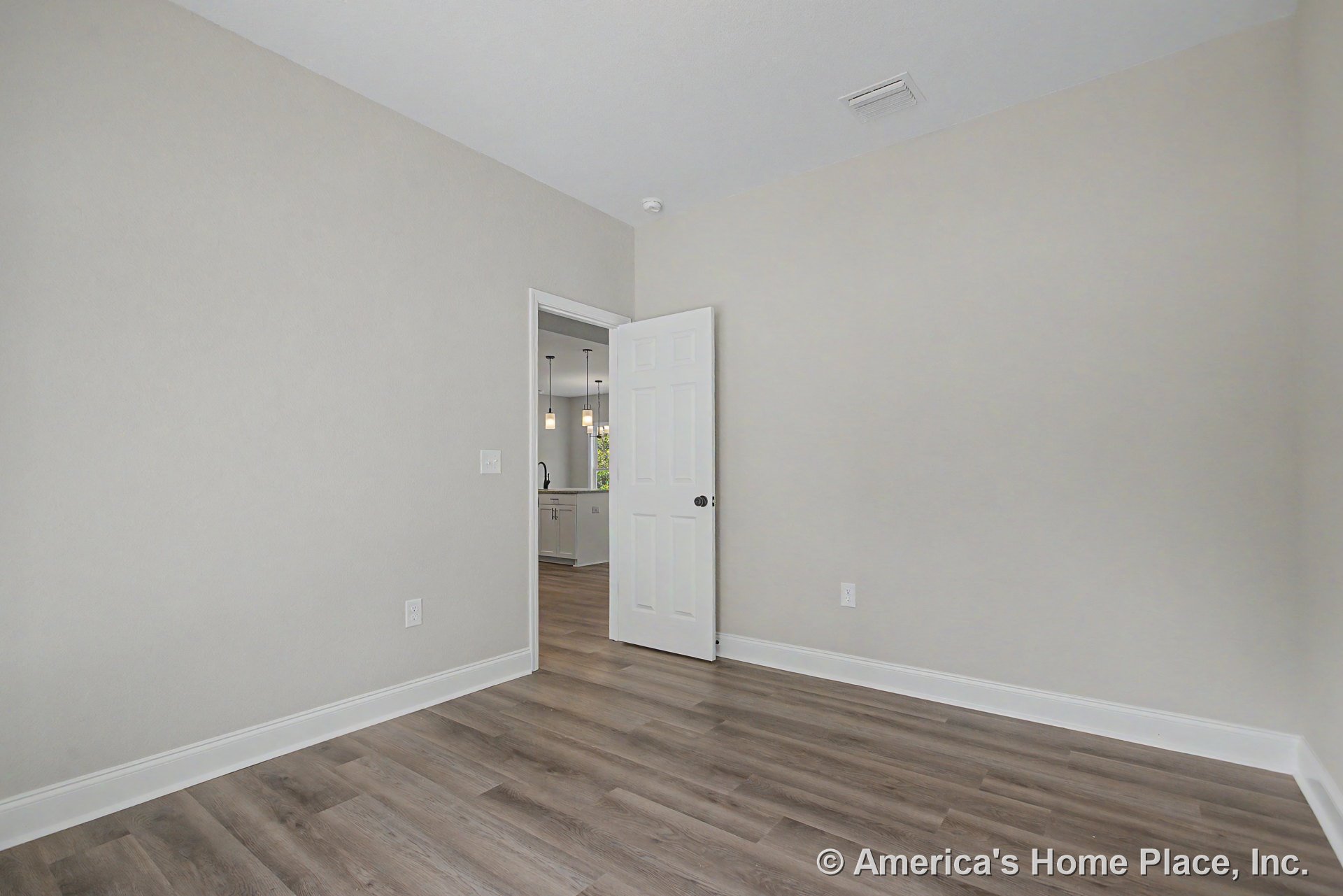 Light wood-look vinyl plank flooring and neutral walls with white baseboard trim, open layout showing white six-panel door, kitchen area with pendant lighting and cabinetry