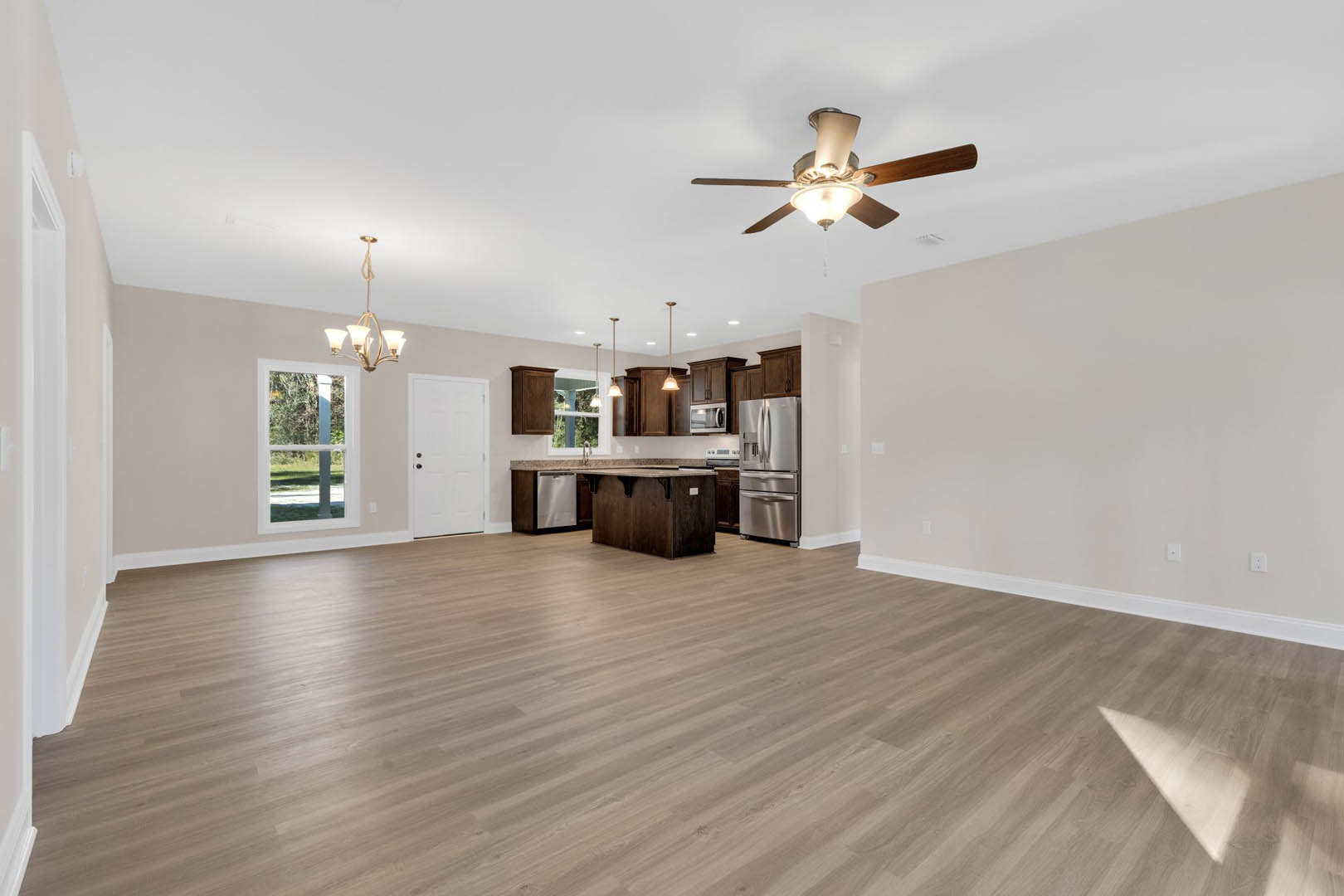Spacious open floor plan featuring a central kitchen island, wood flooring, ceiling fan with light, stainless steel refrigerator, white-framed window, and white door with black