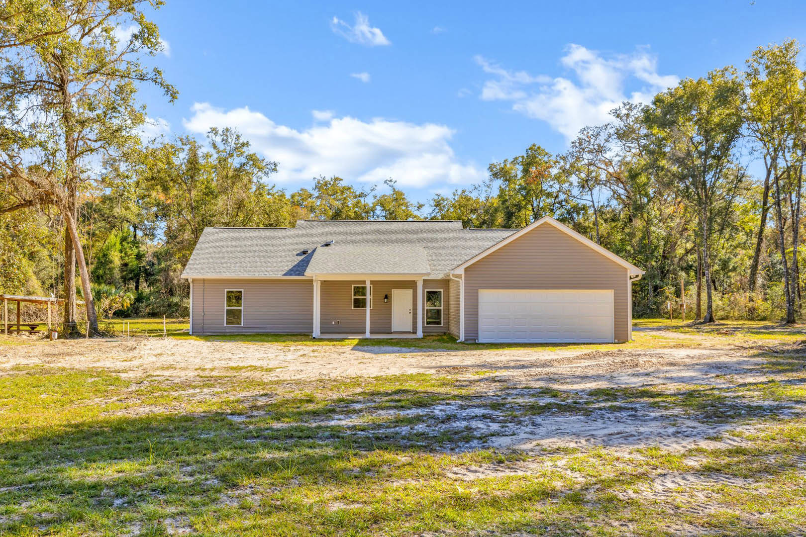 Two-story house with white garage door, light fixture above, gray siding, pitched roof, and mature trees in the background; muddy driveway and patchy grass in foreground.