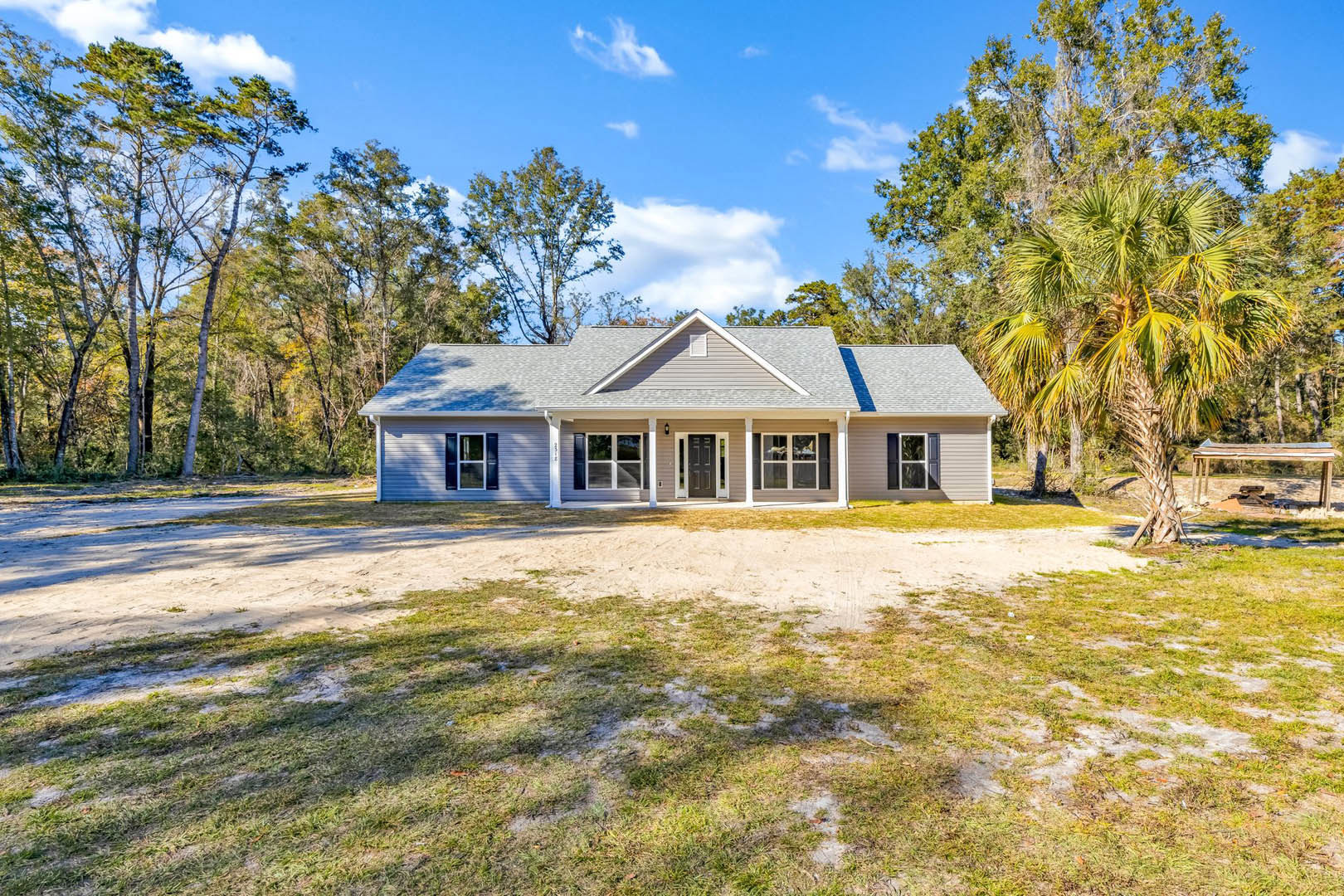 Two-story farmhouse with white siding, covered porch, dirt driveway, palm tree, and surrounding grassy yard with scattered trees.