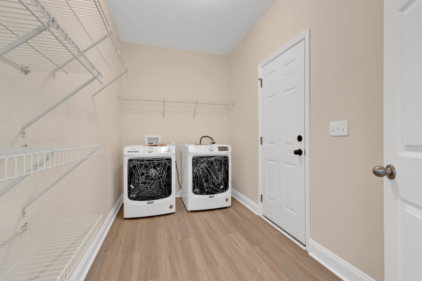 White front-loading washing machine with shattered glass door in a laundry room, white shelving above, light-colored walls, and black door knob visible.