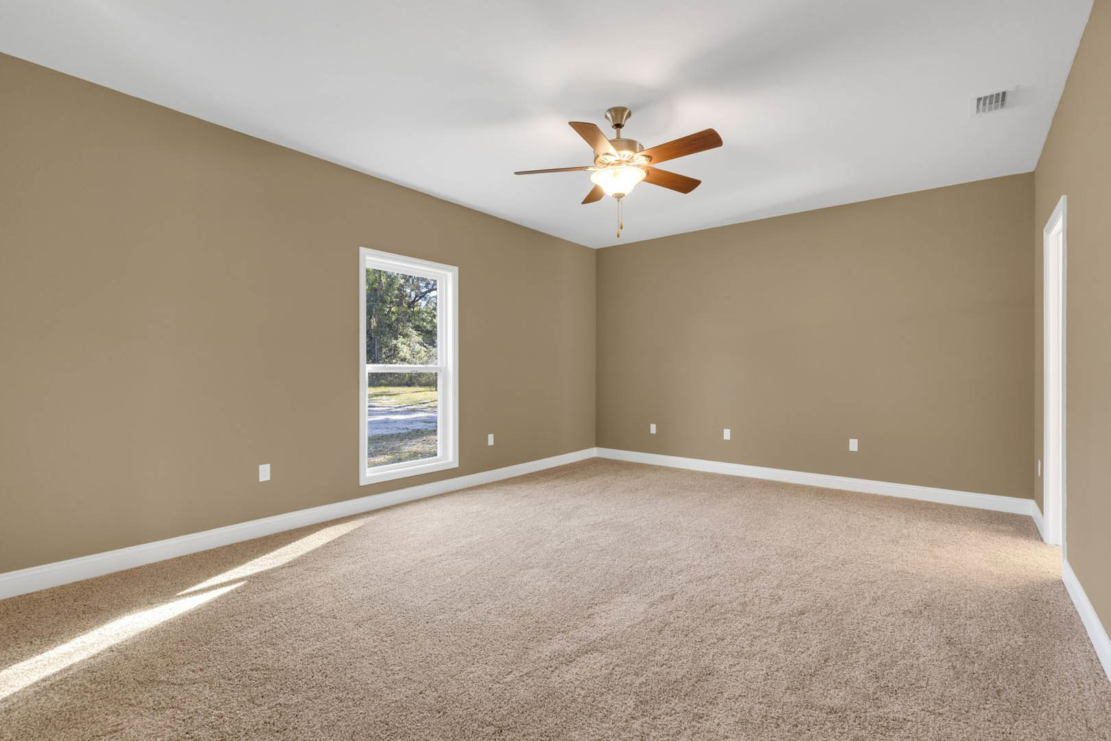 Neutral-toned carpeted room with white walls, ceiling fan with light fixture, large window overlooking green trees, natural light illuminating the floor