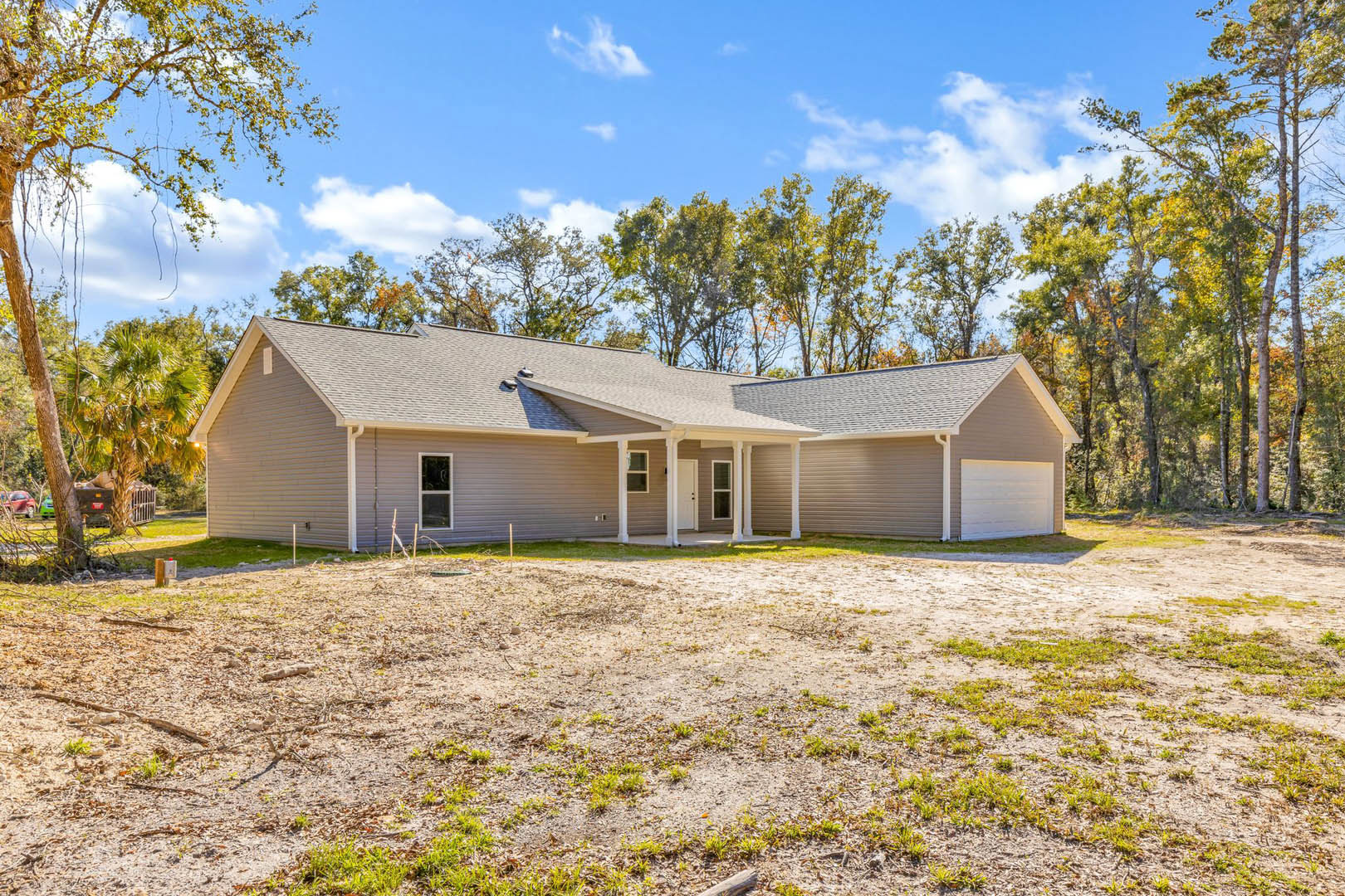 Two-story home with gray siding, white trim, covered front porch, white door, paved driveway, patch of bare dirt, large windows, surrounded by mature trees under cloudy sky