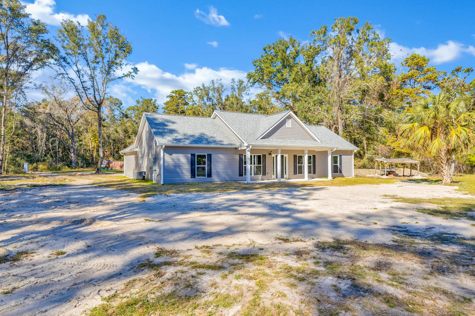 White-roofed farmhouse with large driveway, surrounded by grass and trees, featuring white-framed windows and a small wooden structure with a roof near a palm tree.