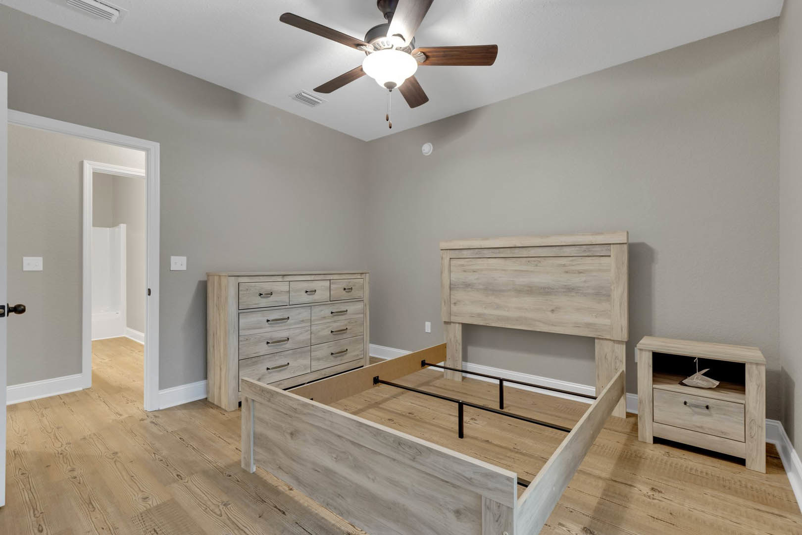 Bedroom with wooden bed frame and storage drawers, ceiling fan with light fixture, wooden nightstand displaying a decorative boat, dresser with drawers and storage box, open door