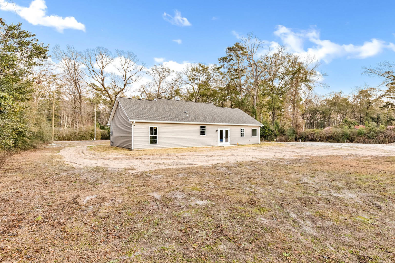 Two-story house with gray roof and white double doors, large windows, surrounded by grass and leafy trees, dirt road and open field in the background under cloudy sky