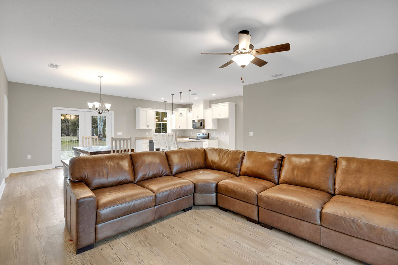 Brown leather couch facing a wooden dining table and chairs in an open living room with light walls, ceiling fan, and hardwood floors