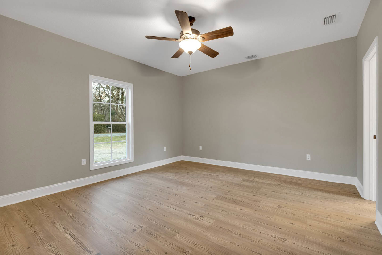 Ceiling fan with light fixture mounted on white ceiling, large window overlooking green yard, wood flooring, wall vent visible