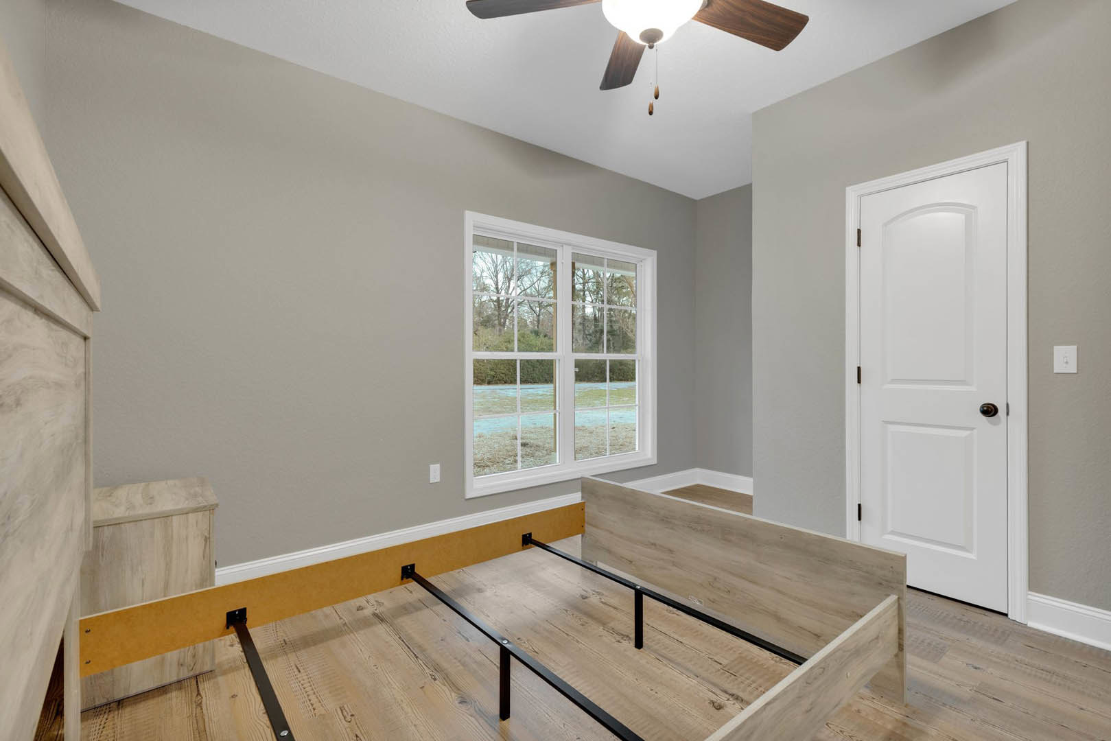 Ceiling fan with light fixture above metal bed frame on wood floor, white door with black handle, window overlooking river, metal railing along wall
