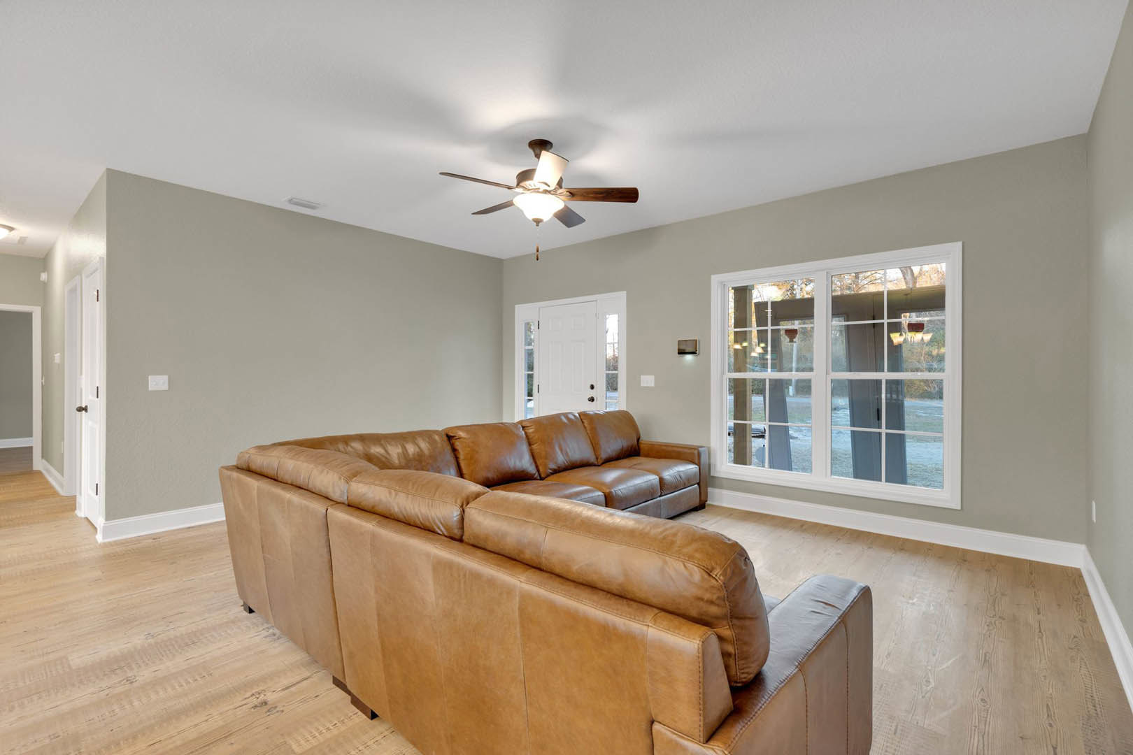 Brown leather couch facing a window, ceiling fan with light fixture overhead, white walls and trim, glass-paneled door, hardwood flooring