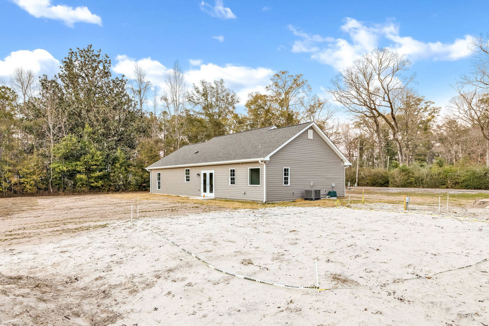 Modern house with white roof surrounded by dirt field, white tape marking ground, leafless tree nearby, and dense forest in background under cloudy sky