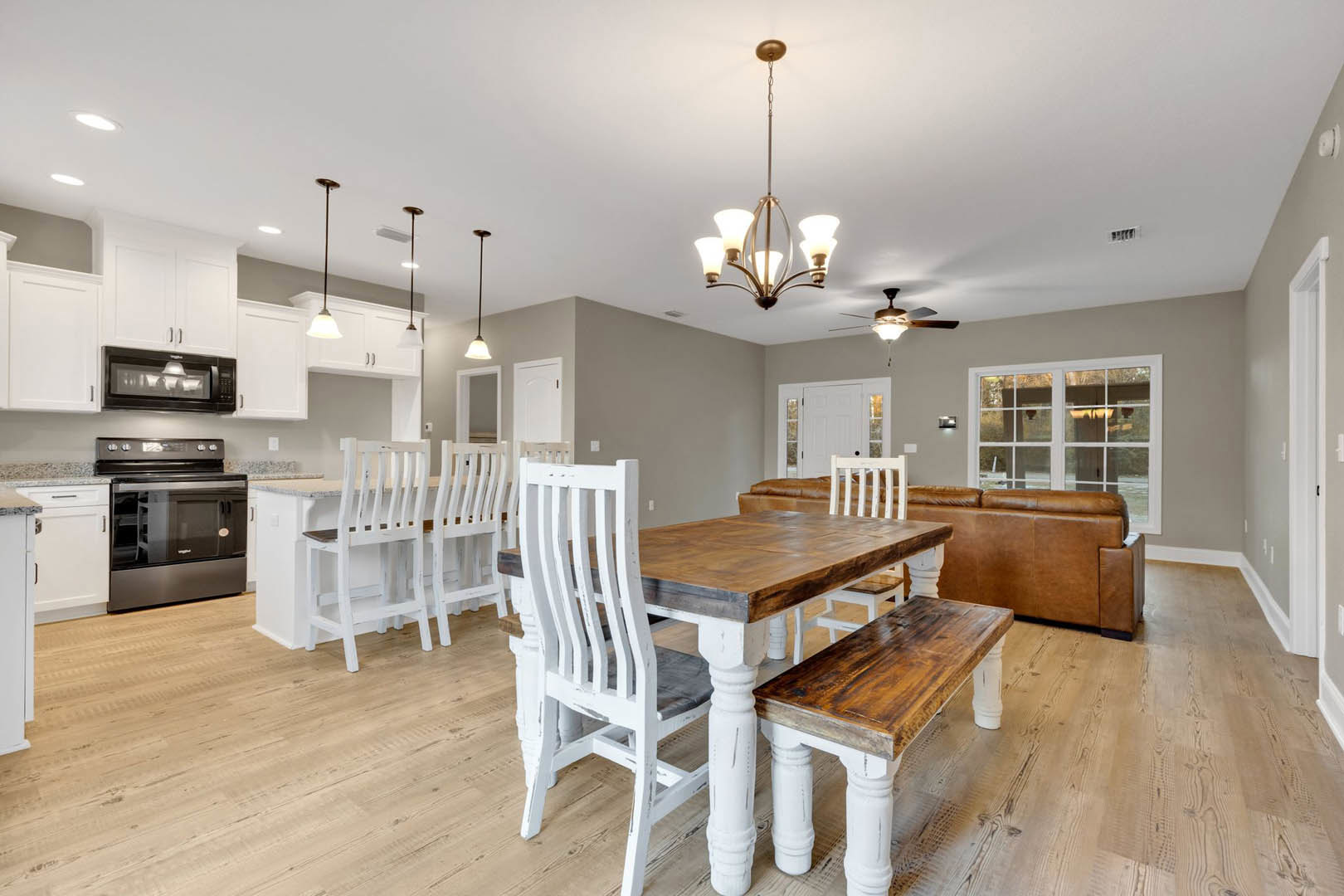 Wood dining table with white chairs set on hardwood floor, open to kitchen with stainless steel stove, white cabinetry, and glass oven, under recessed lighting and neutral walls.