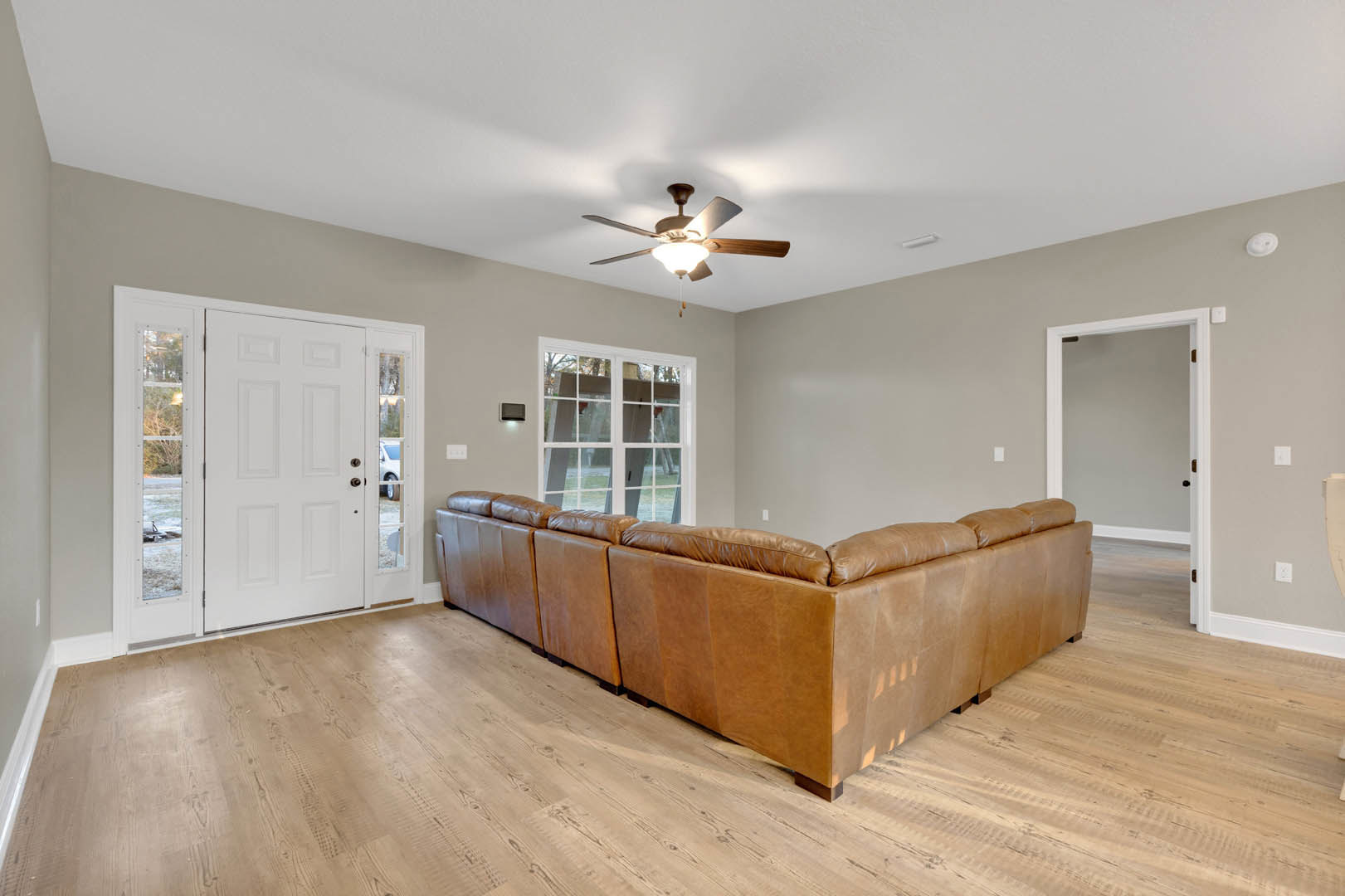 Brown leather couch on laminate wood flooring, ceiling fan with light fixture, white door with glass windows, large window with white trim, neutral walls, modern interior finishes.
