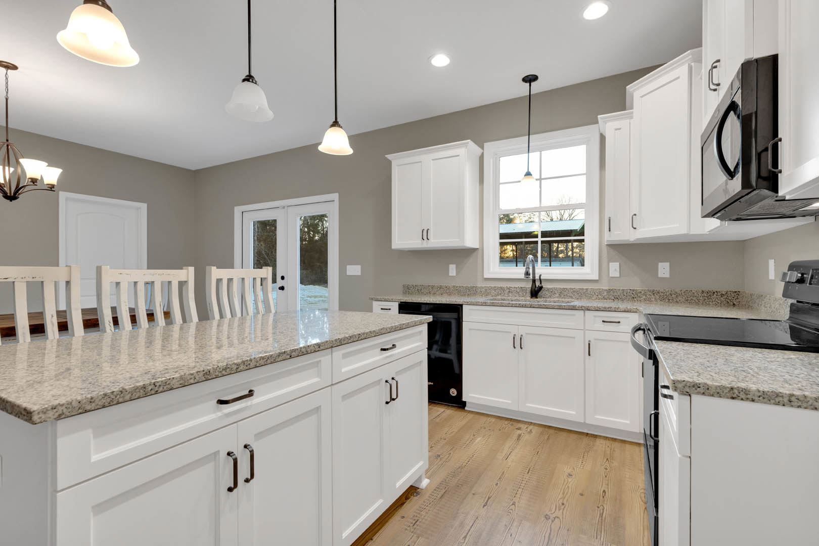 White kitchen with granite countertops, stainless steel appliances, white cabinetry, island with barstools, pendant light fixtures, glass-paneled door, and large window providing