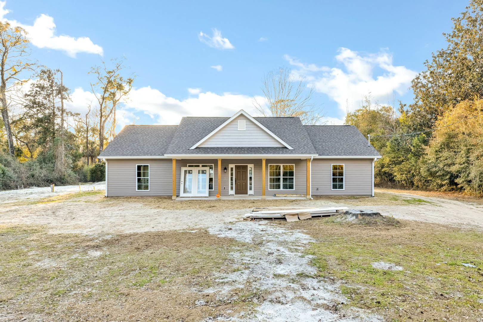 Two-story custom home under construction with exposed framing, brown double front doors with glass panels, white-framed windows, dirt yard, surrounded by mature trees under a clear