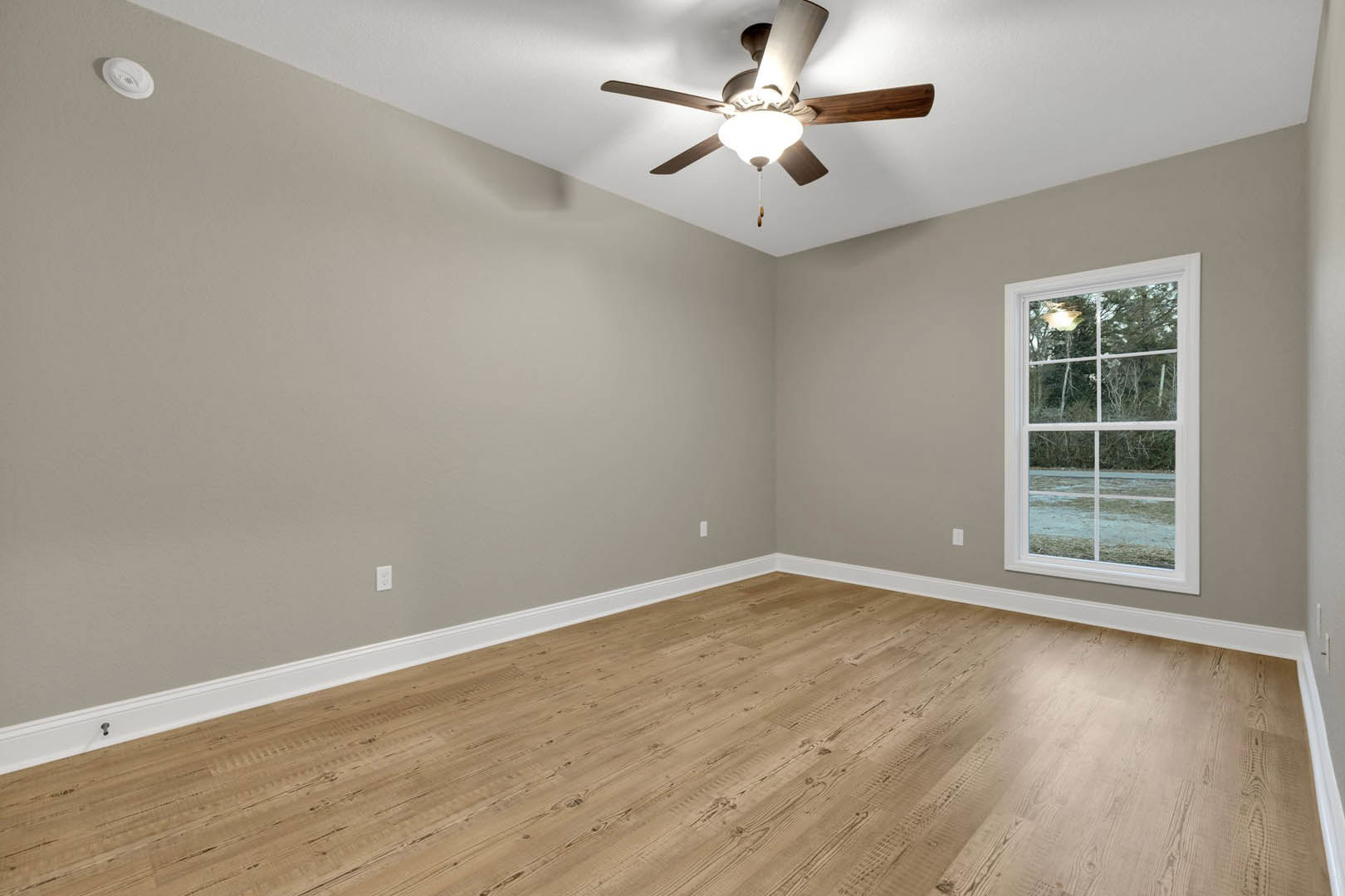 Bedroom with wood flooring, white baseboards, ceiling fan with light fixture, large window overlooking trees, white walls, and recessed lighting.