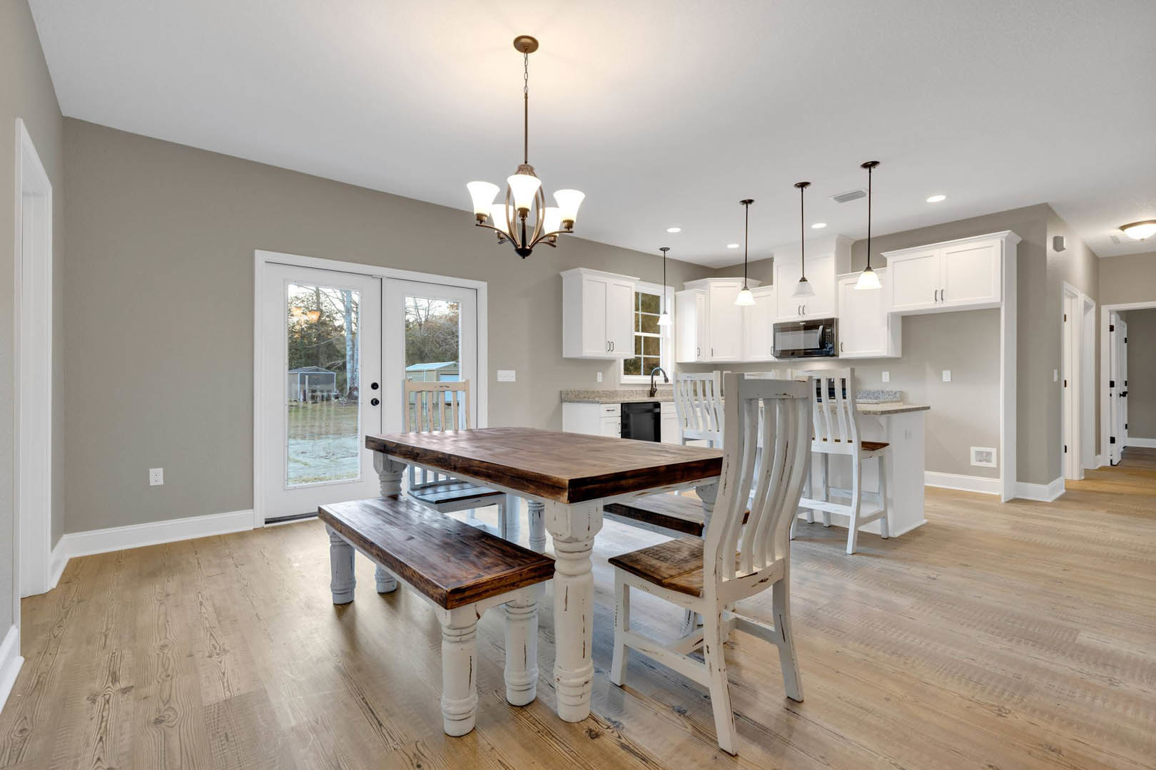 Dining area with wooden table, white chairs, and bench set on laminate flooring, adjacent to kitchen and glass doors
