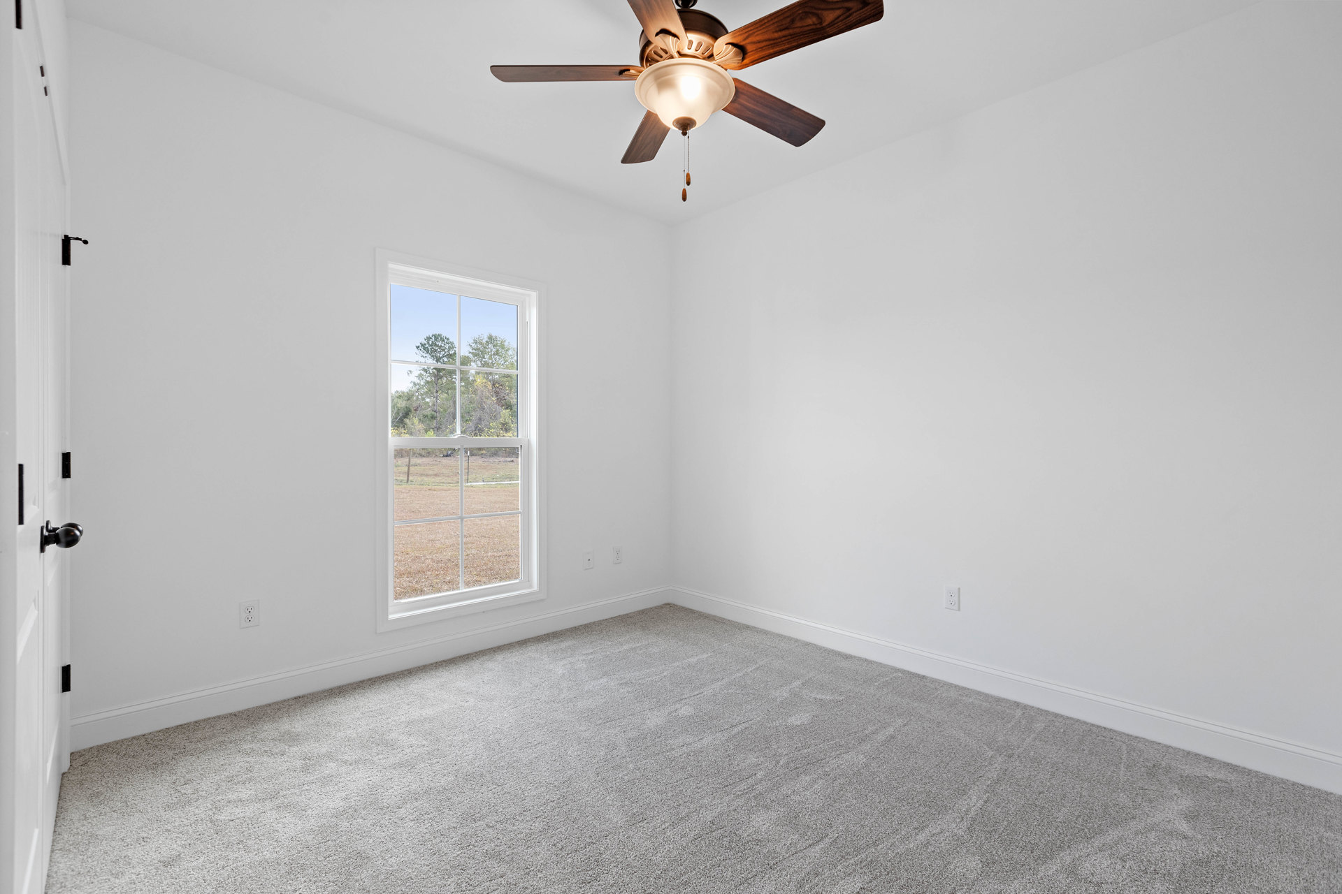 Carpeted bedroom with white walls, ceiling fan with light fixture, large window overlooking grassy field and trees