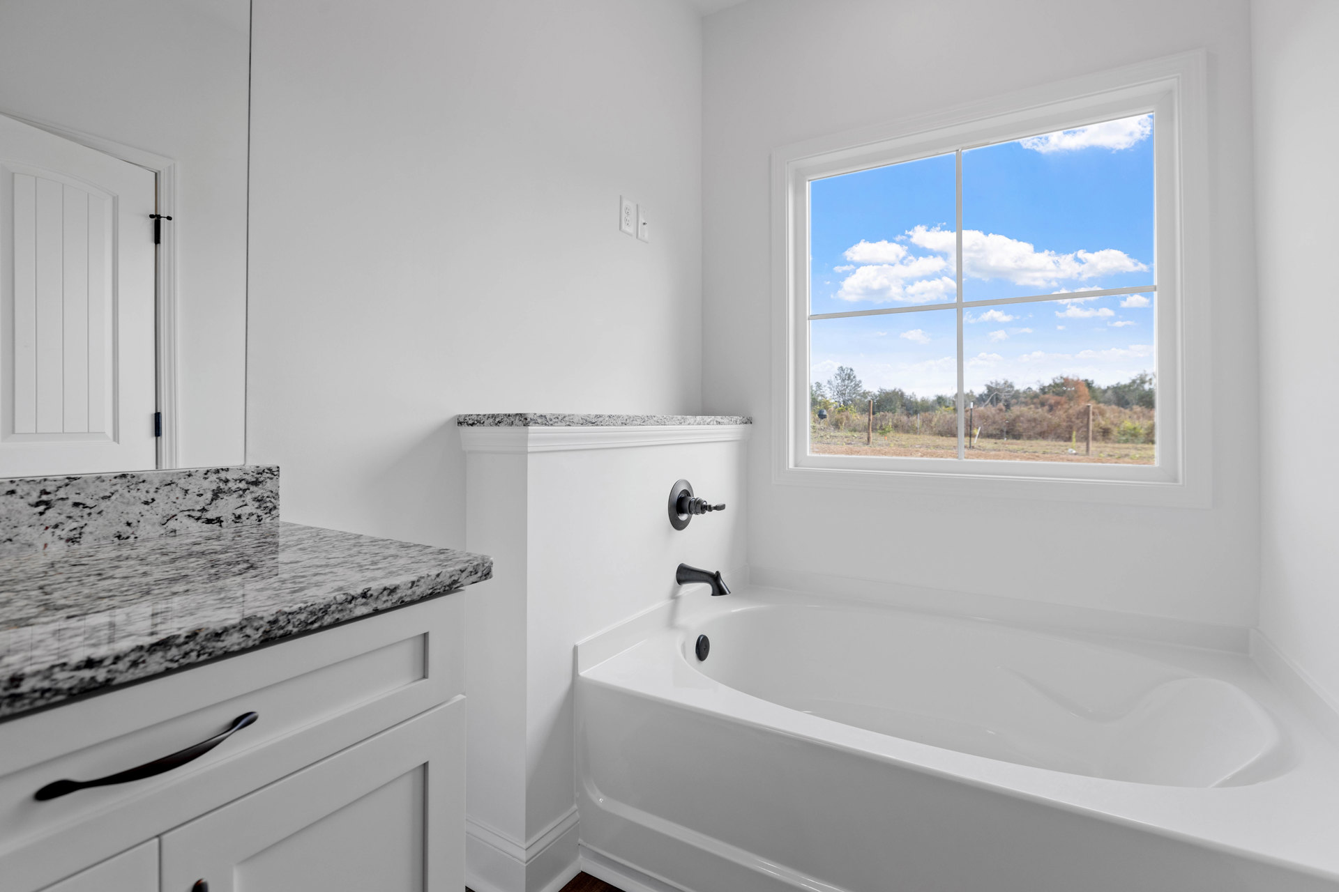 Freestanding white bathtub beneath large window overlooking field and trees, light wood vanity with black hardware, white door with black handle, neutral tile flooring