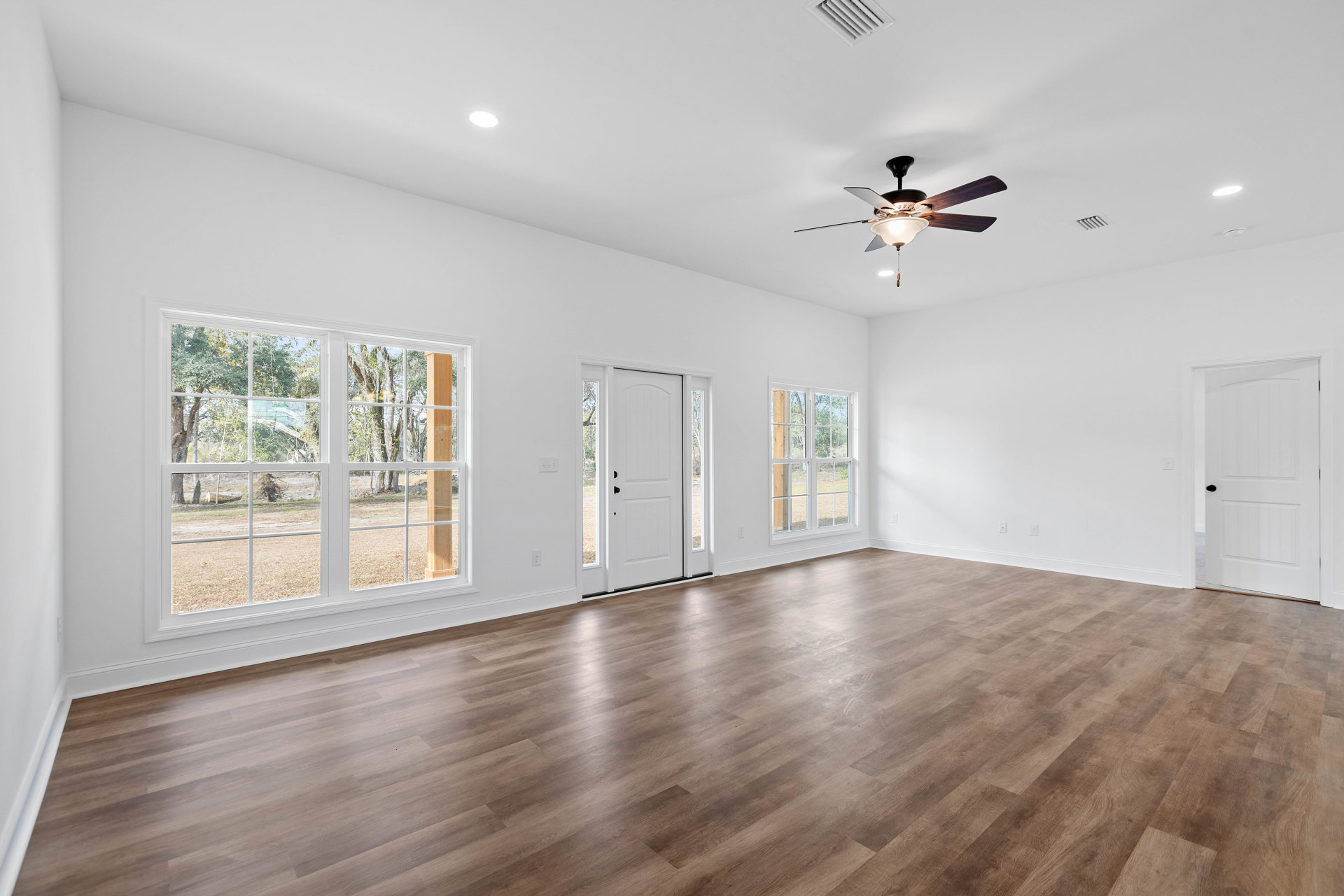 Wood flooring and white walls in a bright room with a ceiling fan, glass-paneled white door, and windows.