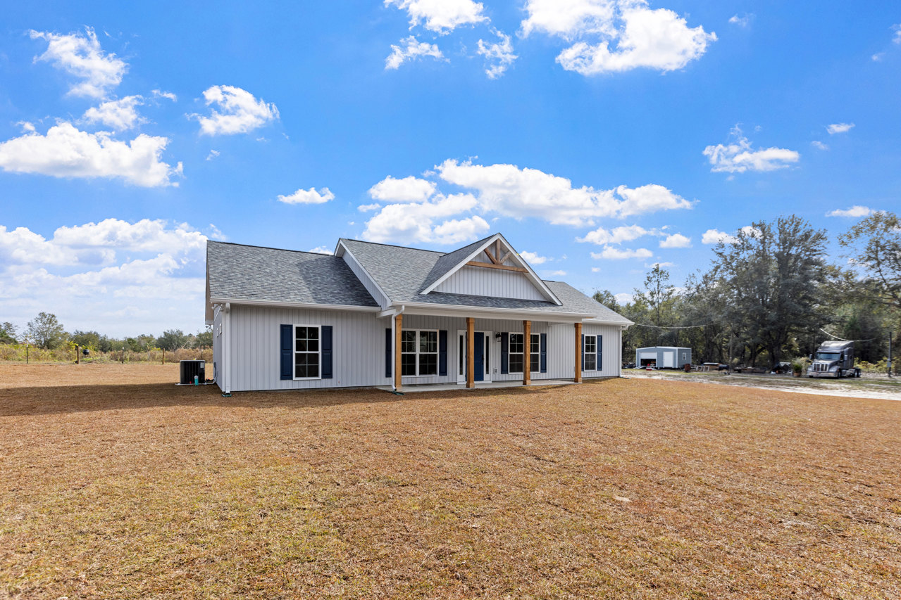 White house with covered porch, gray roof, and multiple windows, set behind a wide expanse of green grass under a blue sky with scattered clouds; pickup truck parked near the road.