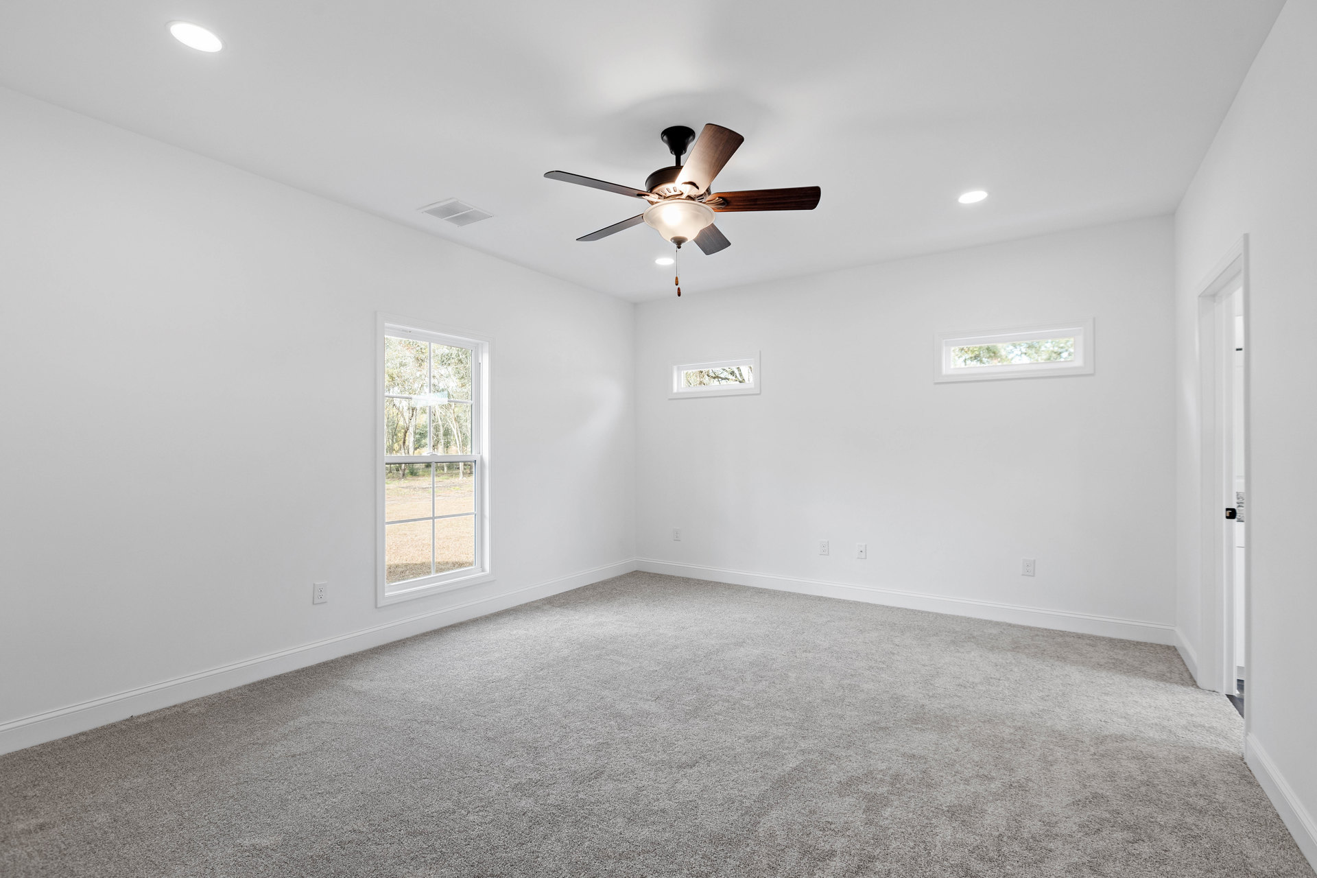 Carpeted room with white walls, ceiling fan with light fixture, large windows showing trees outside