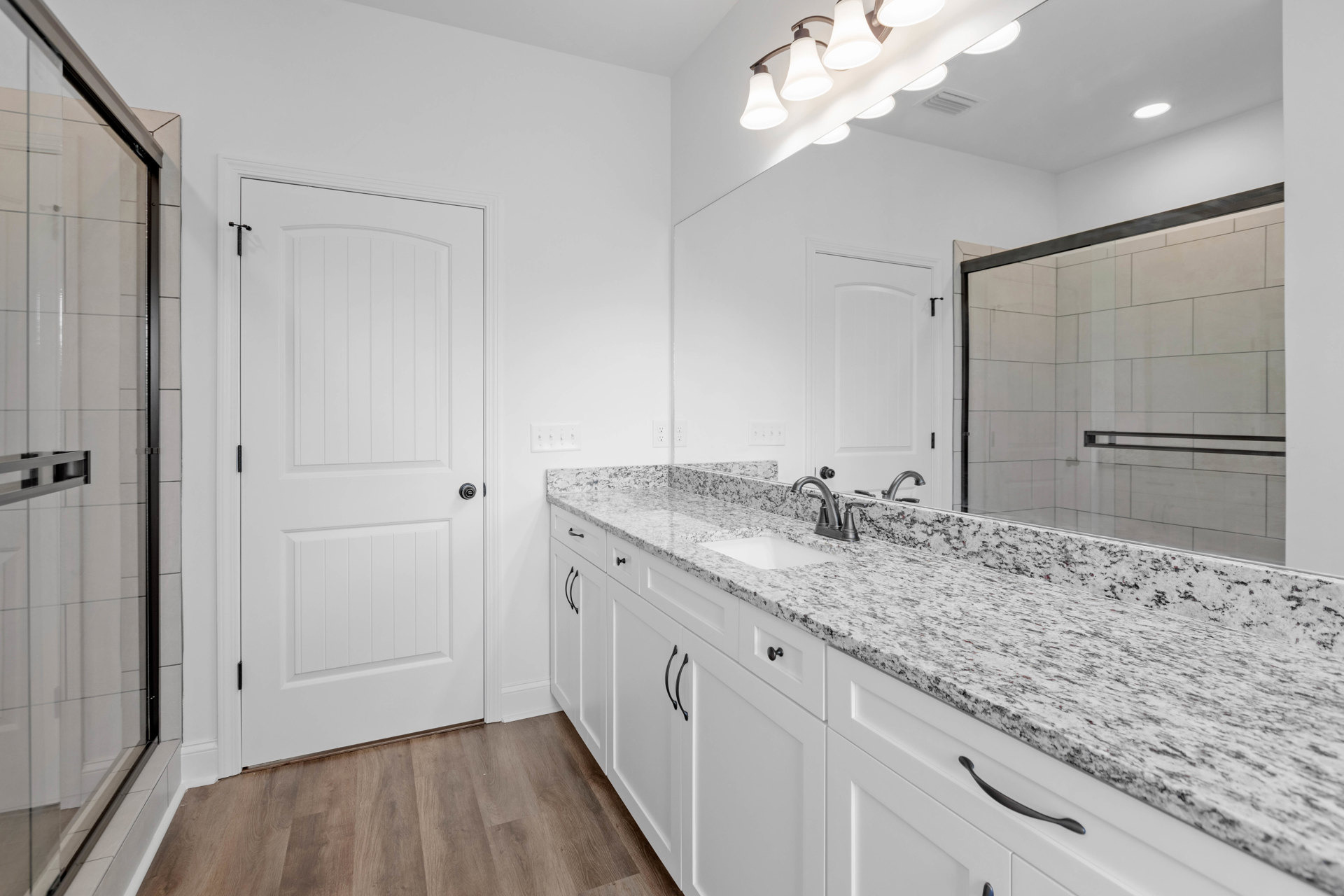 Bathroom with marble countertop, rectangular mirror above sink, glass shower door with black metal bar, white cabinetry with black handles, wood flooring, and white walls.