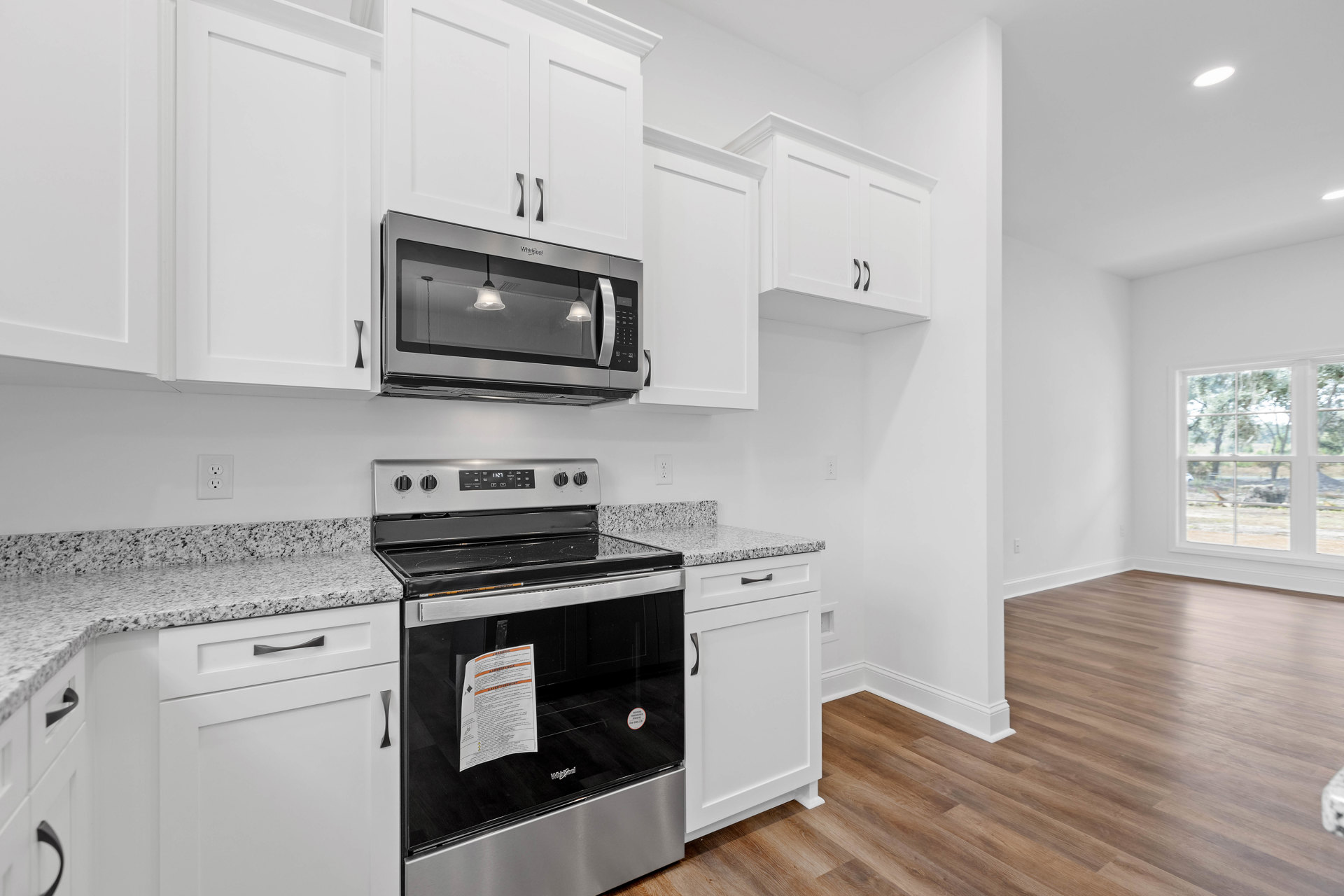White kitchen cabinets, built-in microwave above stove with paper on cooktop, recessed ceiling lights, window showing trees, white door with black handle
