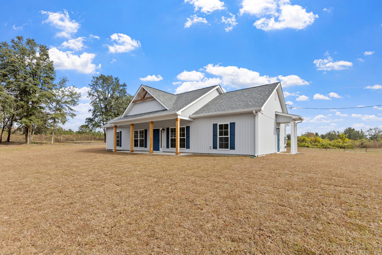 Two-story house with blue roof and blue shuttered windows, surrounded by a large yard with brown grass, leafy tree, and partly cloudy sky